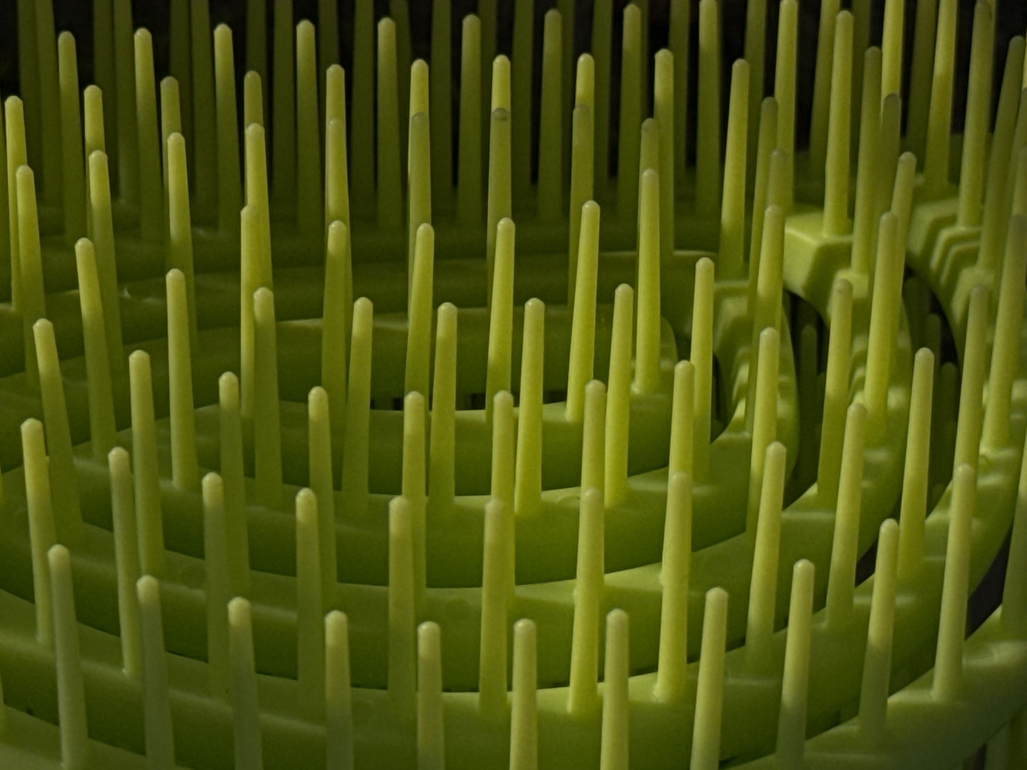 The image is a close-up, abstract shot of a bright green, plastic object with numerous parallel prongs extending horizontally. The prongs are arranged in layers and appear to be part of a larger structure, possibly a comb or brush, with a darker background that creates a strong contrast and depth of field. The lighting highlights the texture and rounded ends of the prongs. There are no people, actions, or interactions visible. The location is not specified in Moscow, Russia, or anywhere else.