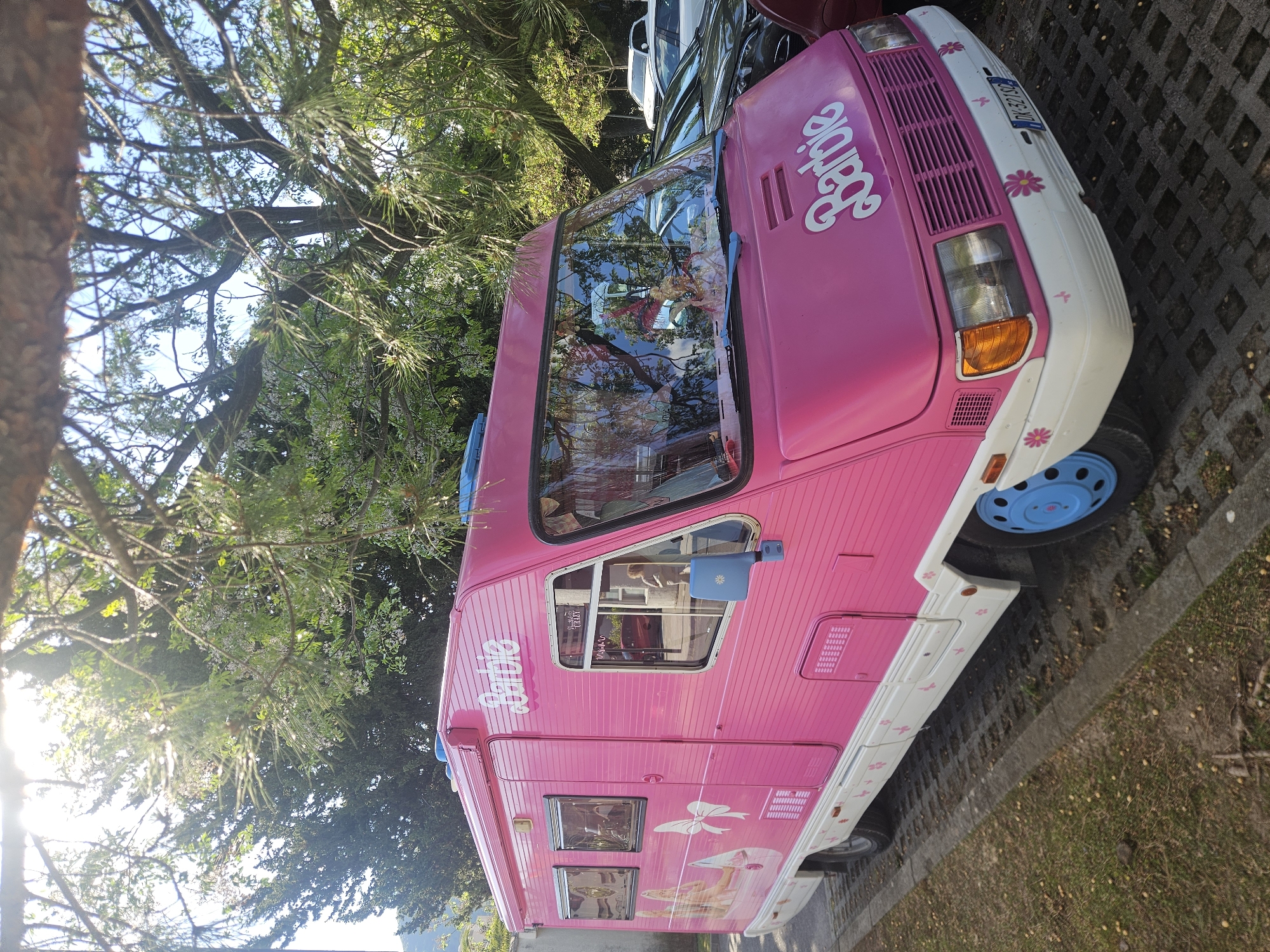 The image shows a pink Barbie-themed camper van parked in an area paved with rectangular stone blocks, adjacent to a grassy area with scattered leaves. The van is decorated with the word "Barbie" on the side and front, and features flower designs on its white trim. The wheels are blue. The camper is parked near trees; the sun is visible through the trees to the left of the frame. The license plate of the vehicle is visible. Other cars are parked nearby. The image orientation is vertical. The location is likely Italy.
