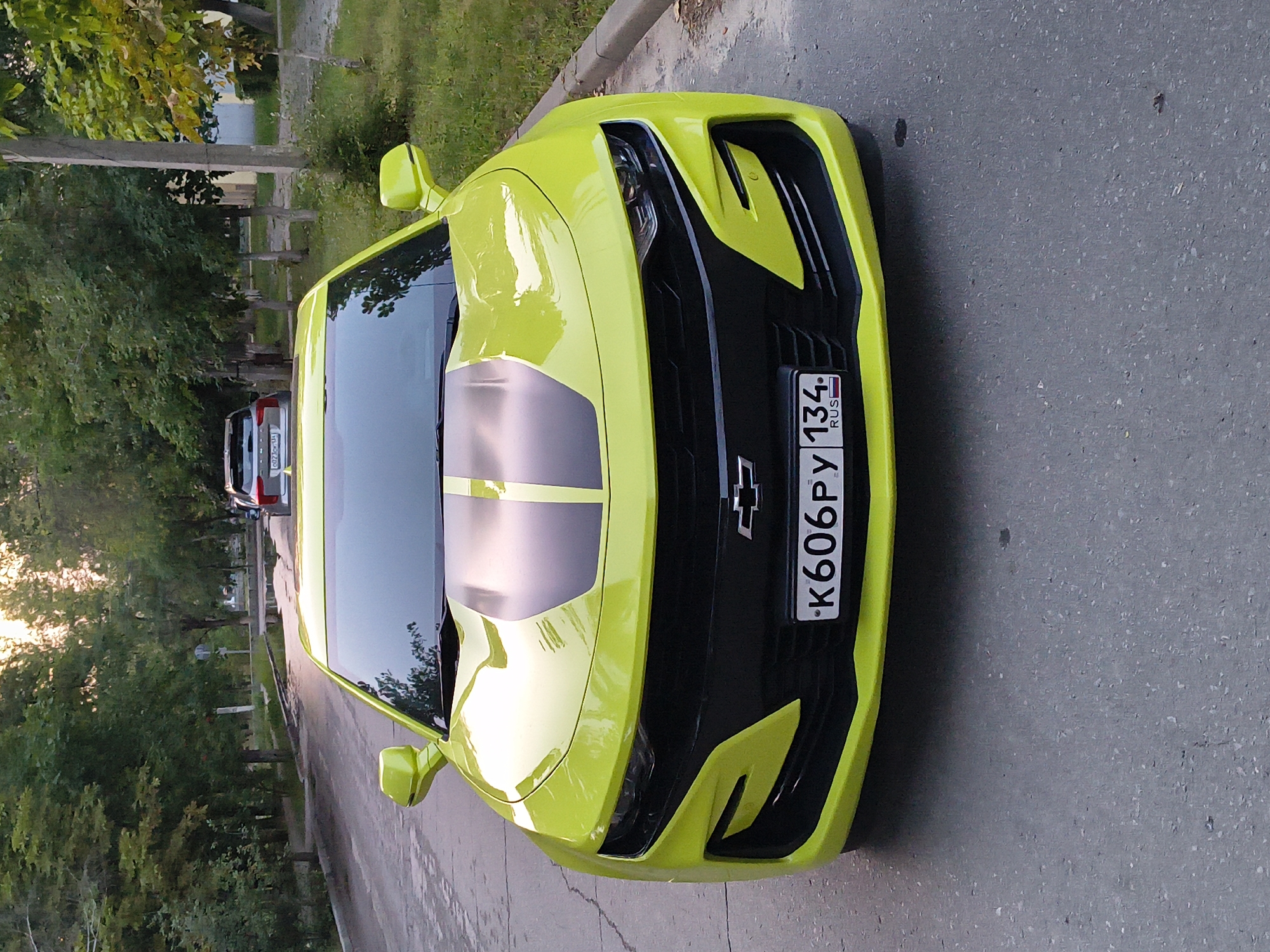 A bright lime green Chevrolet Camaro is parked on an asphalt road in Mikhaylovka, Russia. The car is facing the camera, with its headlights and grille clearly visible. The car has black racing stripes on its hood. The license plate reads "K606PY 134 RUS". In the background, there are trees lining the street, and another car is parked further down the road. The sky is overcast with the sun shining through, creating a soft light. The scene appears to be a residential street.