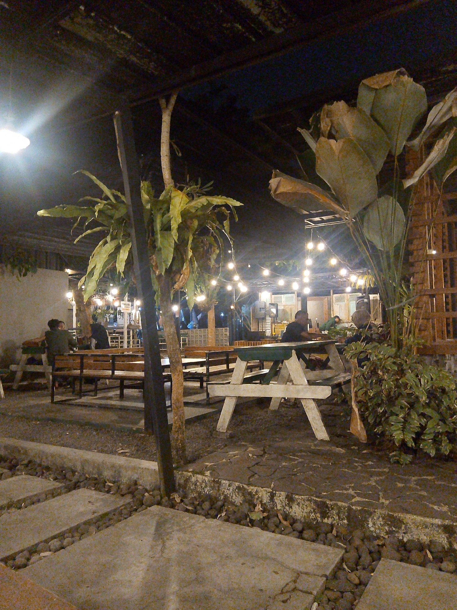 The image depicts an outdoor seating area of a restaurant or cafe at night, likely in Tasikmalaya, Indonesia, given the context. The scene is illuminated by string lights and a bright overhead lamp, casting a warm glow on the surroundings. Several picnic tables with benches are arranged on a gravel-covered ground, interspersed with concrete pathways.
In the foreground, concrete slabs are laid out as a path, bordered by small stones. A tall, dark vertical post stands prominently in the center, part of the overhead shelter structure. To the left, a large fern plant hangs from a tree, its fronds extending outwards. Further back, a cluster of large, light-colored plants with broad leaves dominates the right side of the frame. The background reveals more seating areas, wooden walls, and a glimpse of what appears to be an interior space.
Several individuals are present in the image, seated at the tables, engaged in conversation or dining. Two people are visible at a table on the left, facing away from the camera. Further back, several more figures are seated at tables, their forms silhouetted against the ambient light. One person is seen looking towards the camera at a table in the mid-ground. The overall atmosphere suggests a relaxed, social gathering place.