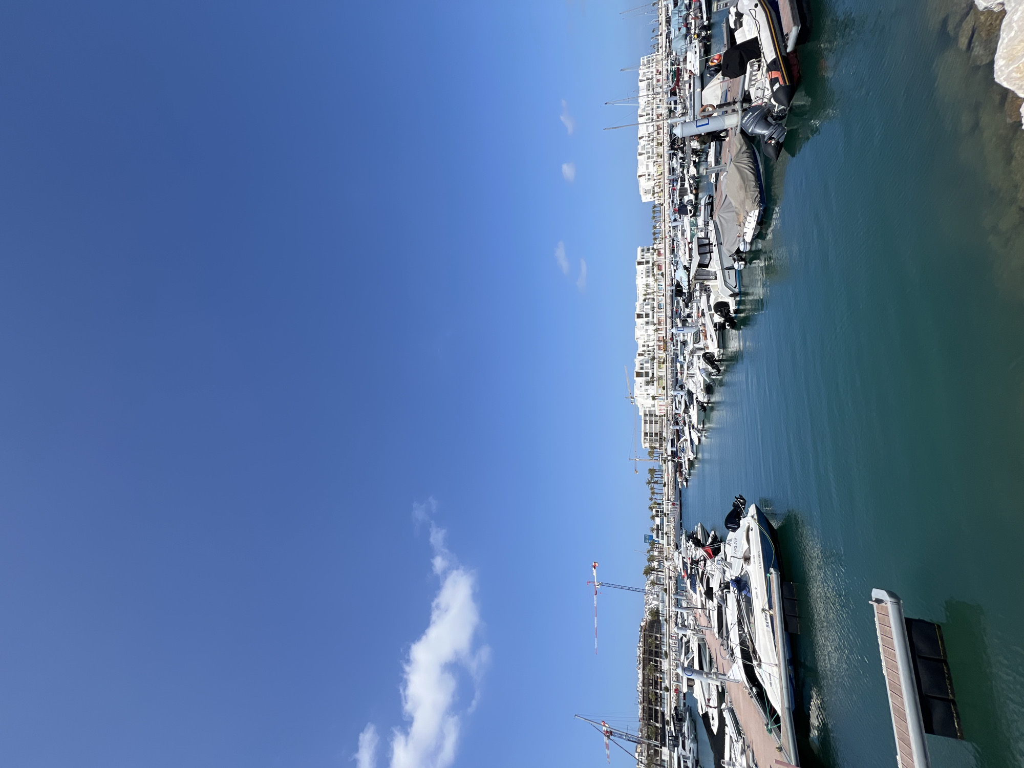 The image displays a marina in Tanger, Morocco, under a clear, bright blue sky with sparse white cumulus clouds in the upper left quadrant. The scene is dominated by a dense arrangement of numerous leisure boats, including motorboats and rigid inflatable boats, moored along grey floating docks. The water is a deep teal color, reflecting the surrounding environment. Most boats are white, featuring outboard motors (some with visible "YAMAHA" branding) and some covered with dark tarpaulins. One visible boat registration is "7-YG-219" with coordinates N 3 34.098' W 5 53.670'.
In the background, a series of modern, multi-story white buildings with terraces line the waterfront, interspersed with green palm trees. Beyond these structures, at least two prominent red and white construction cranes are visible on the left side, indicating active development in the area. Several small, indistinct figures are visible on the docks and near the boats, one in a dark silhouette appears to be standing beside a moored vessel. The immediate foreground on the right shows light-colored rocks at the water's edge. The overall lighting suggests a sunny day.