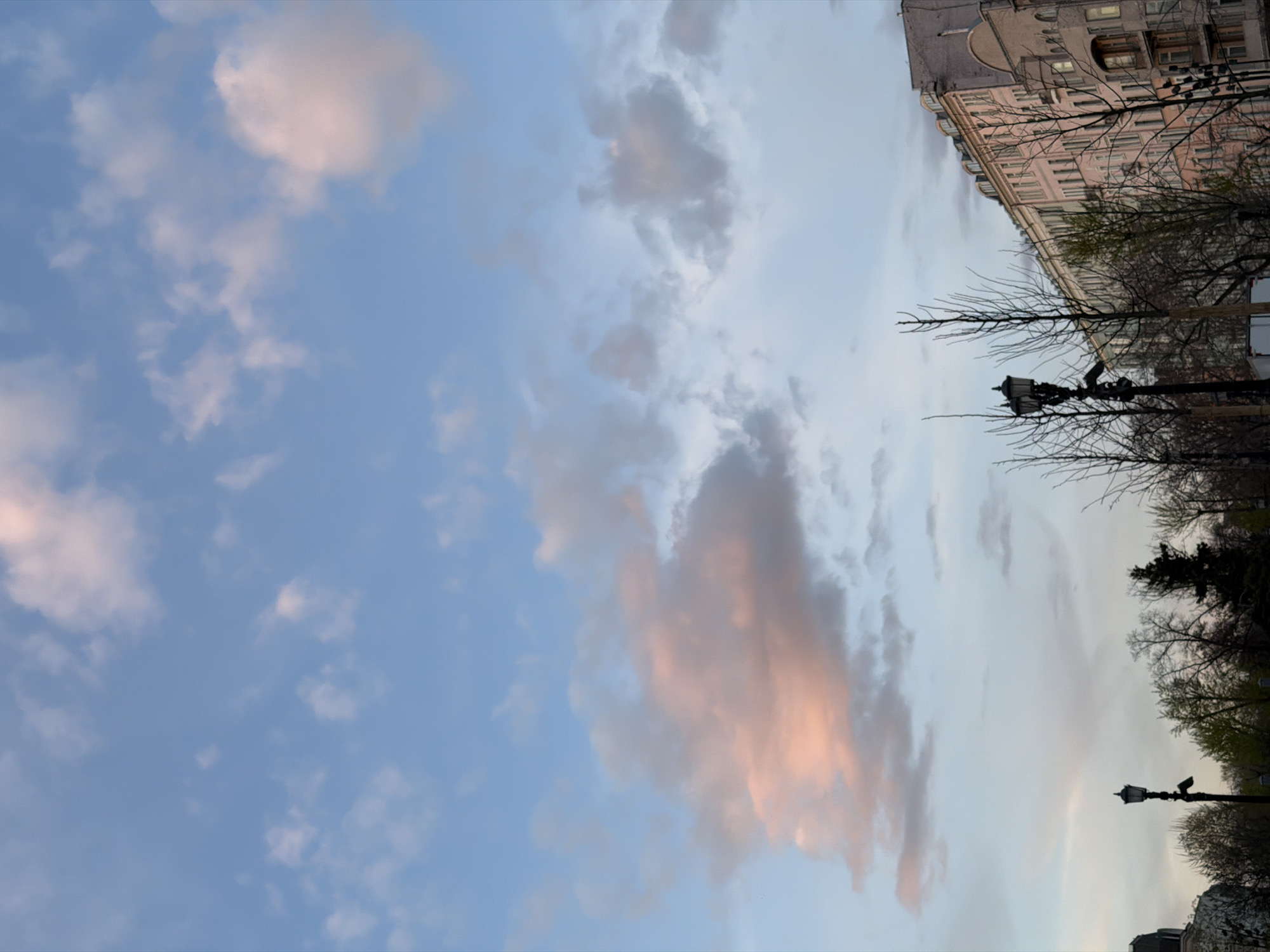 The image shows a view of the sky and a portion of a cityscape. In the sky, there are scattered clouds tinged with pink hues, suggesting either sunrise or sunset. A multi-story building with numerous windows occupies the upper right corner of the frame, seemingly a residential or commercial structure. Adjacent to the building, there are trees with bare branches, indicating either late autumn or early spring. Street lamps are visible among the trees. Based on the architectural style and the general environment, it is plausible that this scene is located in Moscow, Russia.
