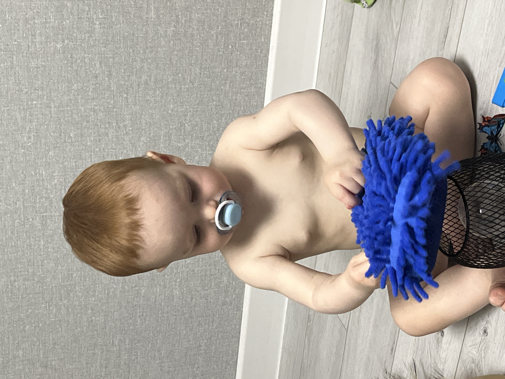 This indoor photograph, taken in Chaikovsky, Russia, features a young child sitting on a light wooden floor. The child has light brown hair and is wearing a light blue and white pacifier. They appear to be topless, with a partial view of a diaper.
The child is actively engaged with a vibrant blue, fluffy object, resembling a duster or a toy, holding it with both hands and looking down at it.
The setting includes a light gray wall with a subtle textured pattern, visible in the background. A white baseboard runs along the bottom of the wall, separating it from the light-colored floorboards, which exhibit a distinct wood grain. To the right of the child, a black mesh basket or container is partially visible, next to a colorful butterfly sticker or decoration (blue, orange, and white) on the floor.
The lighting suggests it is daytime indoors. The child's expression is neutral and focused on the object they are holding. No text is visible in the image.