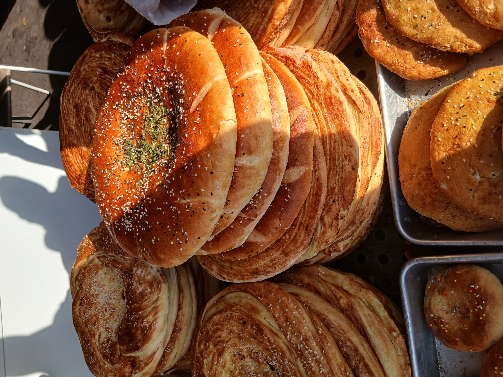 A close-up, overhead shot captures an outdoor market stall in Khankiz, Uzbekistan, laden with freshly baked goods. Several stacks of round, flatbreads dominate the frame. The bread on the left, a lighter golden-brown, features a swirled, layered texture and is sprinkled with white sesame seeds. To its right, a taller stack of similar bread has a deeper brown hue and is adorned with both white and black sesame seeds, along with a small mound of green herbs in the center of the topmost loaf. Further to the right, more bread is visible, some peeking from behind a white plastic bag. In the foreground, two metal trays hold smaller, flatter bread varieties. The tray on the left contains a round bread topped with colorful sprinkles. The tray on the right displays several semi-circular breads, some dusted with sesame seeds. The sunlight casts distinct shadows of people and structures onto a white surface in the upper left corner, suggesting a sunny day at the market.