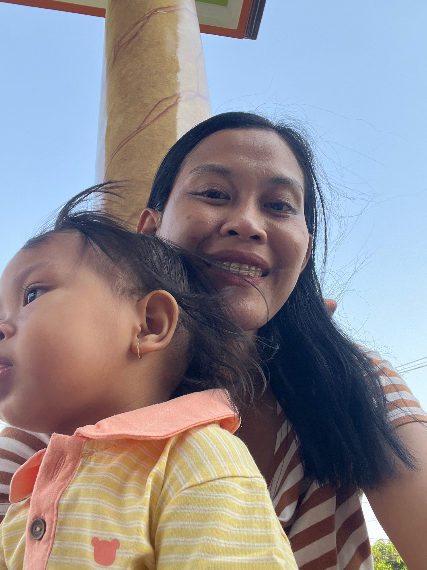 A woman and a young child are captured in a selfie, likely taken outdoors in Kecamatan Pati, Indonesia, as evidenced by the blue sky and a portion of a textured pillar that appears to be part of an outdoor structure. The woman, in the background, has dark hair and is smiling, revealing braces on her teeth. She is wearing a striped shirt. The child, in the foreground and to the left, is looking away from the camera with an open mouth, wearing a yellow and white striped shirt with a coral-colored collar and a small embroidered bear emblem. The child has a small gold hoop earring in their left ear. The wind is blowing, causing strands of hair to fly around the subjects.