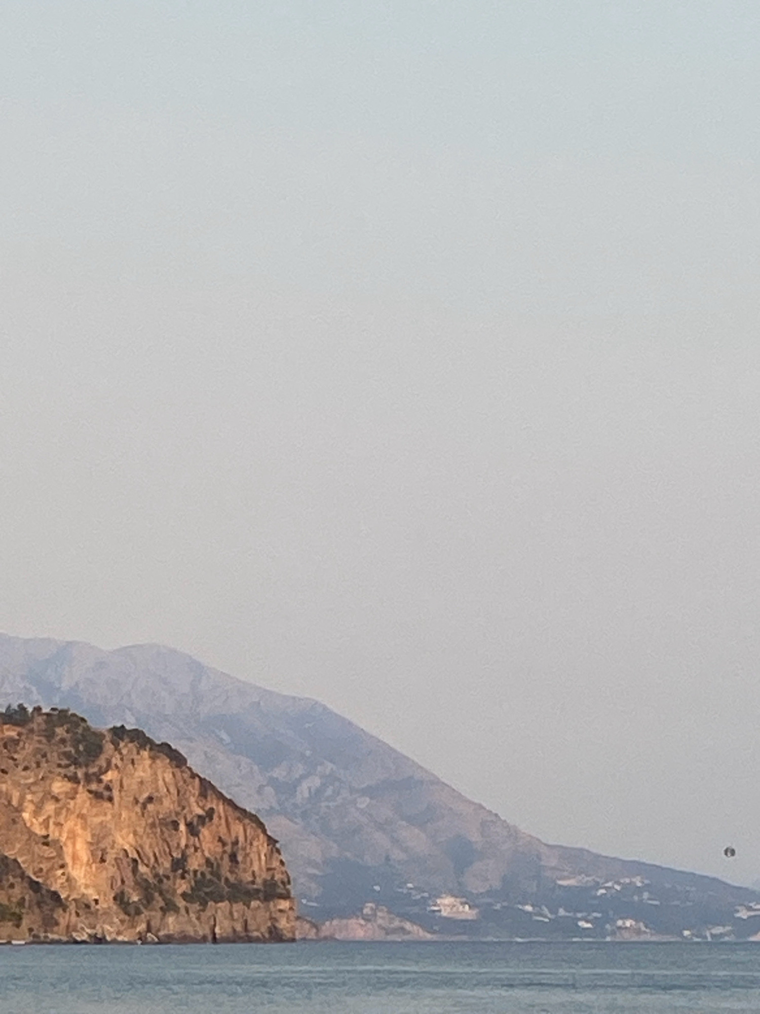 The image depicts a coastal landscape with rugged cliffs and mountains. The foreground features calm, blue water. In the mid-ground, a prominent, sun-drenched cliff face rises from the sea, its texture rough and varied. Behind the cliff, a range of layered mountains recedes into the distance, their peaks softened by atmospheric haze. A small, dark object, possibly a parasailer or a distant bird, is visible in the sky above the mountains on the right side of the frame. The sky is a pale, gradient blue, indicating daylight. The scene is natural and appears to be a view of the coastline near Prijevor, Montenegro.