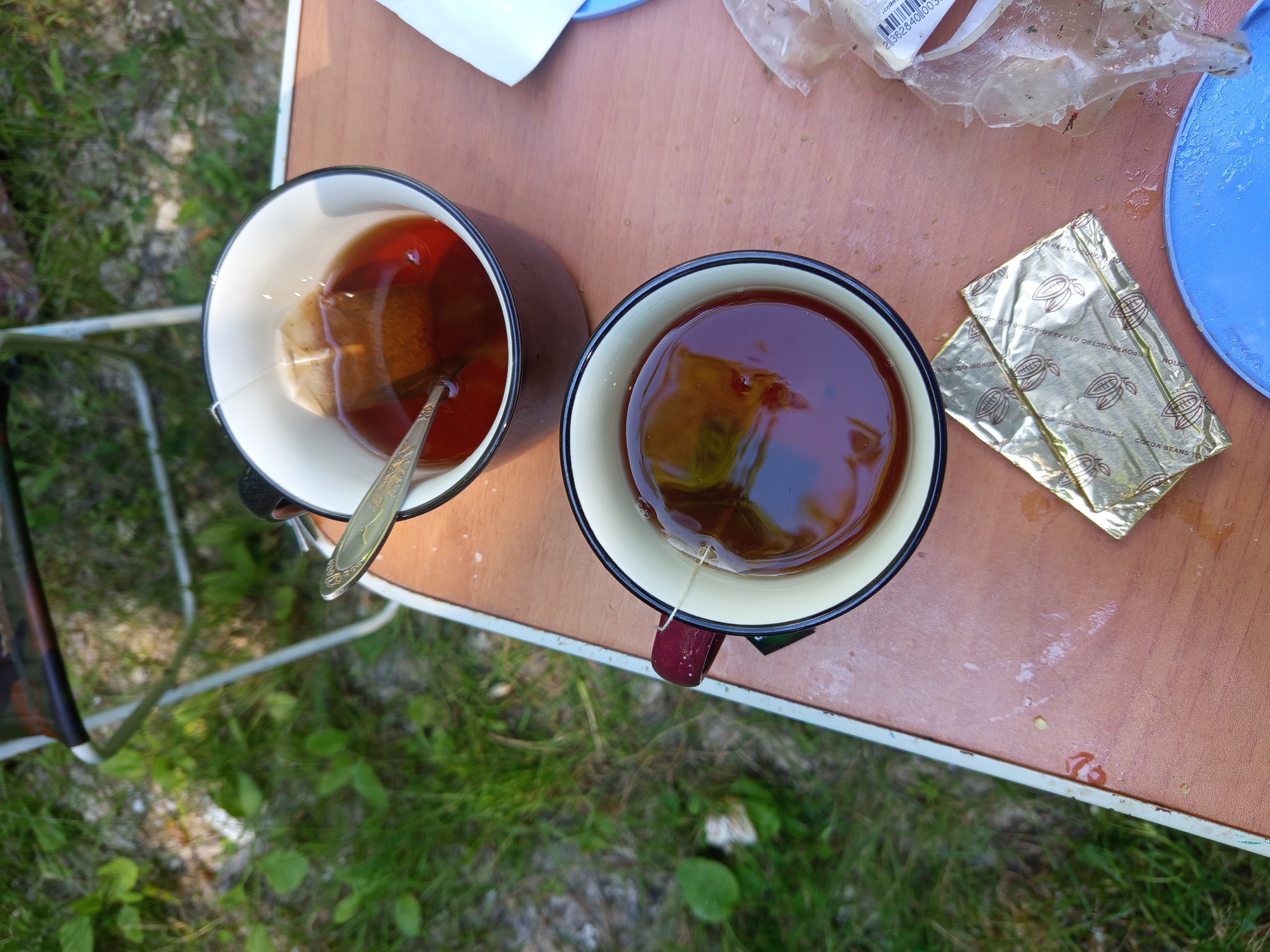 Two mugs of tea are sitting on a wooden table. One mug is in the foreground and has a tea bag in it. The other mug is behind it and also has a tea bag in it, along with a spoon. There are two packets of something shiny and gold, likely wrappers for food items, on the table. A blue plate is also visible. The table appears to be outdoors, as grass and foliage are visible in the background, as well as a portion of a folding chair. The overall scene suggests a casual outdoor setting, possibly a picnic or camping trip, with no specific indication of the city of Giaginskaya, Russia.