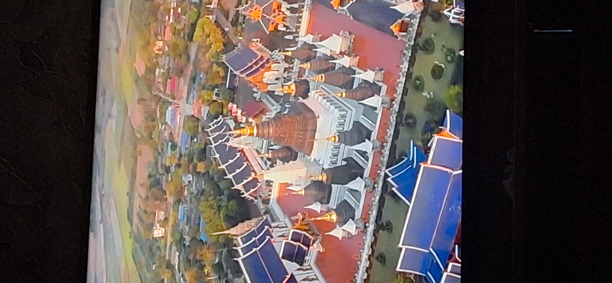 The image shows an aerial view of a temple complex with multiple buildings, stupas, and courtyards. The architecture features prominent blue tiled roofs and white and brown ornate spires. The complex is surrounded by greenery and appears to be located in a rural or semi-rural setting with fields and some scattered houses in the background. The image does not show any people, actions, or interactions. The location is not Bila Tserkva, Ukraine.