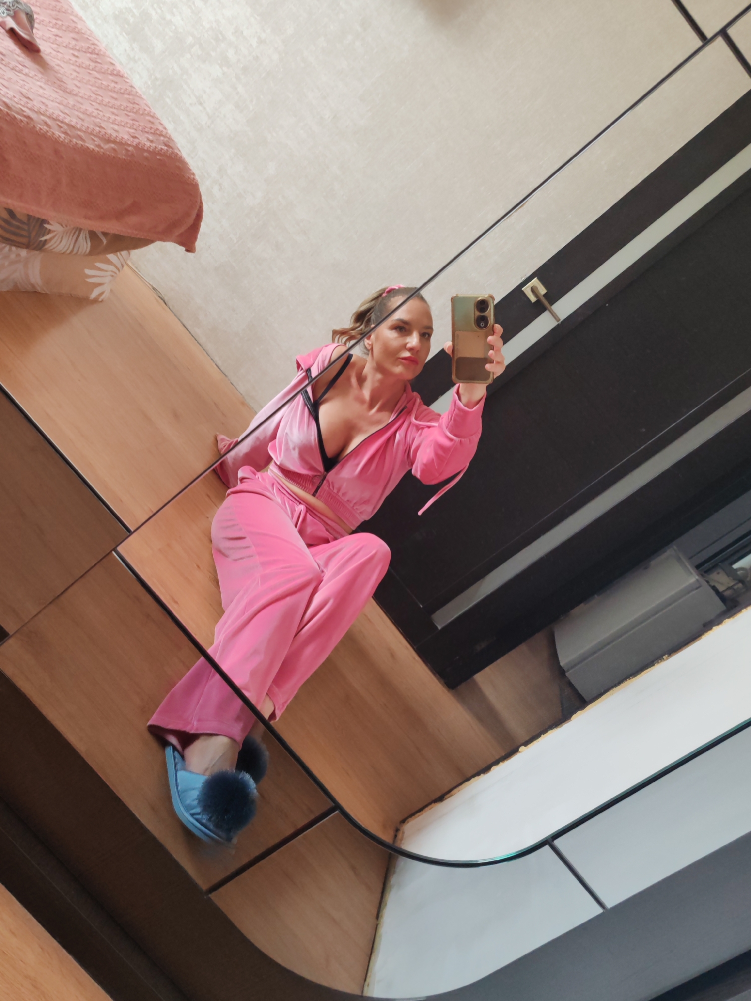 A woman in a pink velour tracksuit takes a selfie in a room. She is wearing blue slippers with fuzzy pom-poms. The room contains a bed with a pink comforter and a wooden floor. The background includes striped wall paneling. There is no indication of the location of the city of Petropavlovsk-Kamchatsky, Russia.