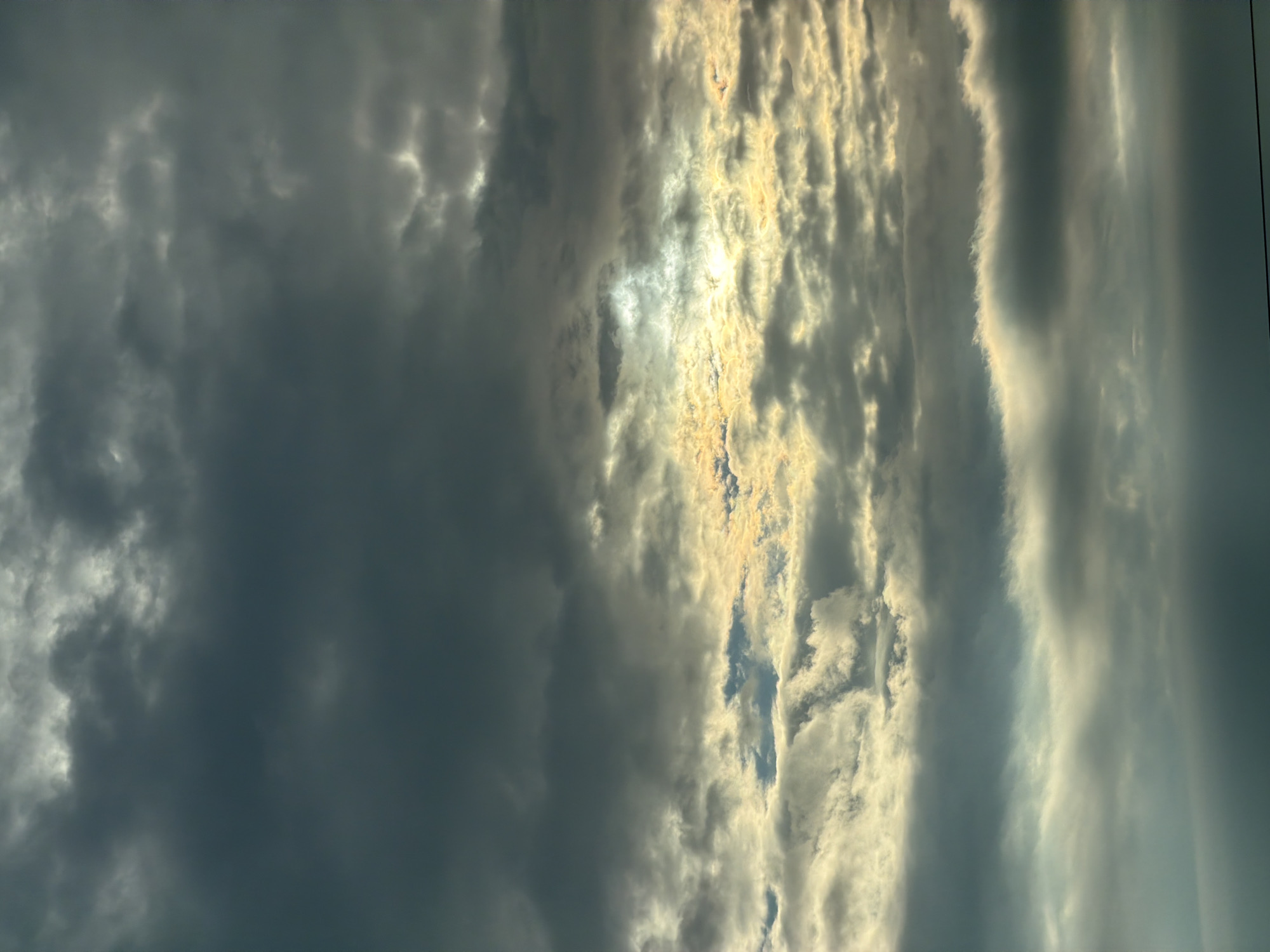 The image captures a dramatic sky filled with layered clouds. Dark, heavy clouds dominate the upper portion of the frame, gradually giving way to lighter, more textured clouds as the eye moves down. Towards the center, a break in the clouds allows sunlight to stream through, illuminating the edges of the surrounding clouds with a golden hue. The lighting creates a sense of depth and contrast, with pockets of light interspersed with deep shadows. The bottom of the image shows a blur of darker clouds, with a faint horizontal line visible at the very bottom, suggesting a distant horizon or possibly power lines. There are no people, subjects, objects, actions, or interactions visible in the frame, as the image focuses solely on the atmospheric conditions of the sky. The location is noted as Unknown, Unknown, indicating that specific geographical information is not available.