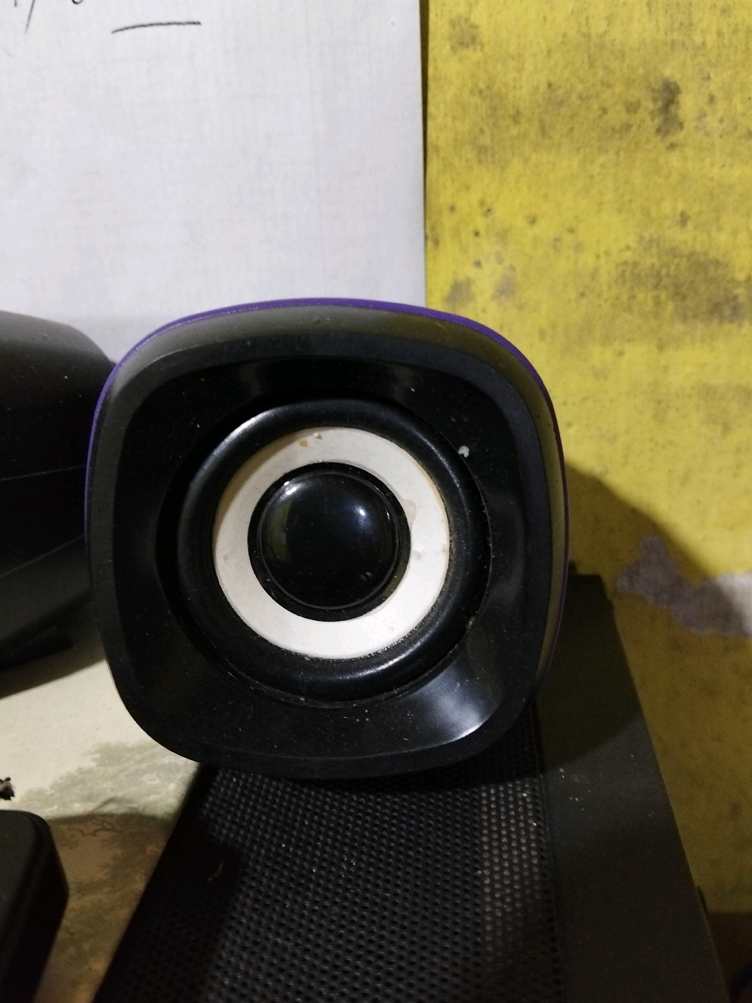 The image shows a close-up of a black computer speaker with a purple accent around the cone. The speaker cone is white with a black center. The speaker is placed on a black perforated surface, likely another speaker or a desk. In the background, there is a white sheet of paper with some faint writing and a yellow textured surface with dark stains. To the left of the main speaker, a portion of another black speaker is visible. The lighting is dim, casting shadows. The scene appears to be indoors, possibly in a home or office setting.