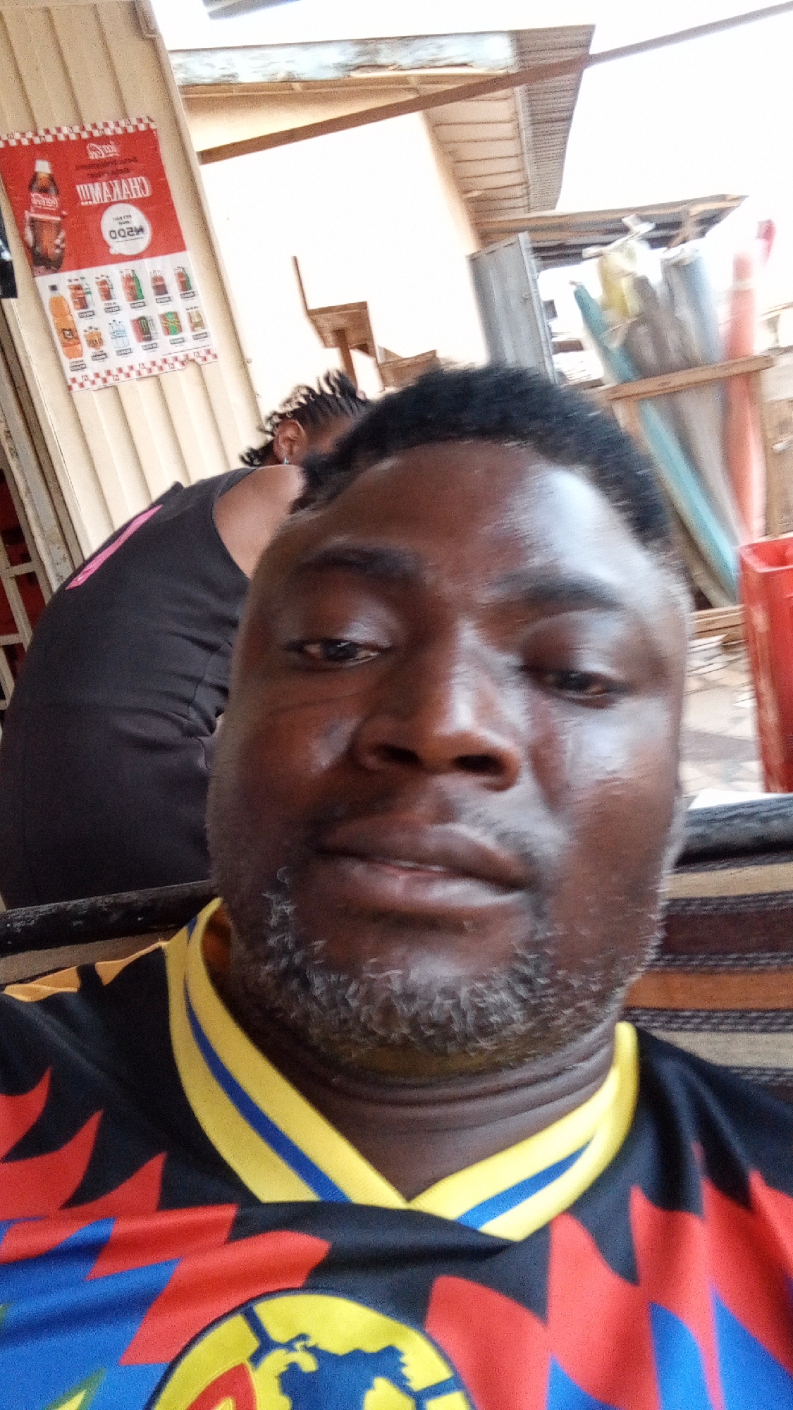 This close-up selfie captures a man with dark skin, short, curly black hair, and a distinct salt-and-pepper beard. He is wearing a black sports jersey featuring a prominent yellow and blue striped collar. The jersey's body is adorned with vibrant red, yellow, and blue geometric patterns, and a circular logo resembling the Club América crest, depicting a globe and a football, is visible on his chest. His gaze is directed towards the camera, conveying a neutral to slightly serious expression.
Behind him, to the left, another individual is partially visible, seen from the back and side, wearing a black garment with a pink stripe and having braided hair. The background indicates an outdoor or semi-enclosed setting, likely a shop or market stall in Sabon Tasha, Nigeria, under bright daytime lighting.
On the left side of the frame, a red and white advertising poster is prominently displayed on a light-colored corrugated metal wall. Due to the selfie format, the text on the poster is mirrored, showing "CHAKAHAN" and "M200" (likely reflecting "NAKAHAC" and "002W" respectively), alongside a visible Coca-Cola bottle and an assortment of other bottled beverages. Further to the right in the background, a bright red plastic bin or container is noticeable, along with various items, possibly tools or merchandise, stored in an open wooden structure.