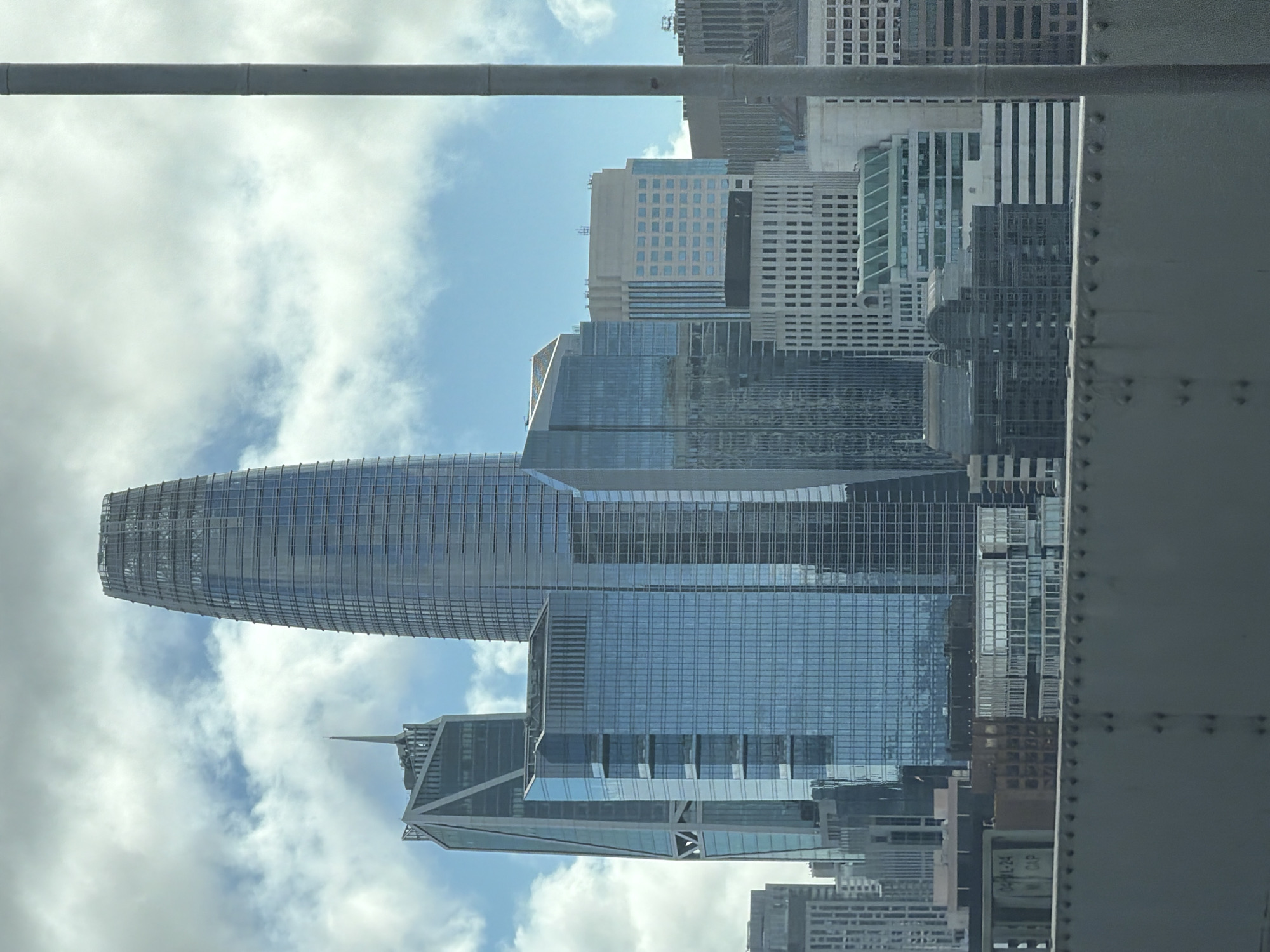The image shows a daytime, exterior, medium shot of the San Francisco skyline, featuring several modern skyscrapers. The Salesforce Tower is the most prominent structure, a tall, slender building with a distinctive tapered top, dominating the center of the frame. To its left is another skyscraper with a sharp, angular design and a spire. Various other buildings of different heights and architectural styles, primarily composed of glass and steel, fill the background. The sky is partly cloudy with patches of blue sky visible. In the foreground, a grey, metallic structure, possibly a bridge railing, runs horizontally across the bottom of the image, partially obscuring some of the lower buildings. A vertical grey pole is visible on the right side of the frame, also in the foreground. No people are visible in the image.
