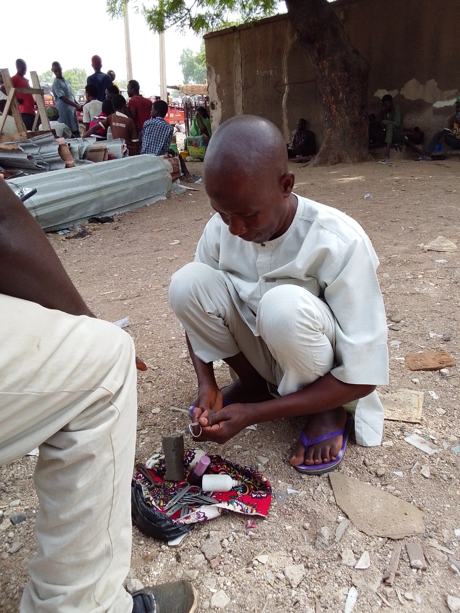 In Jimeta, Nigeria, a man squats outdoors on a dirt ground, examining a ring with tools. He wears a light-colored outfit and purple sandals. Beside him, another person's legs, clad in light pants and sneakers, are visible. Around the man are scattered tools, fabrics, and small containers. In the background, blurred figures gather near a structure, and some individuals are seated under a tree near a building.