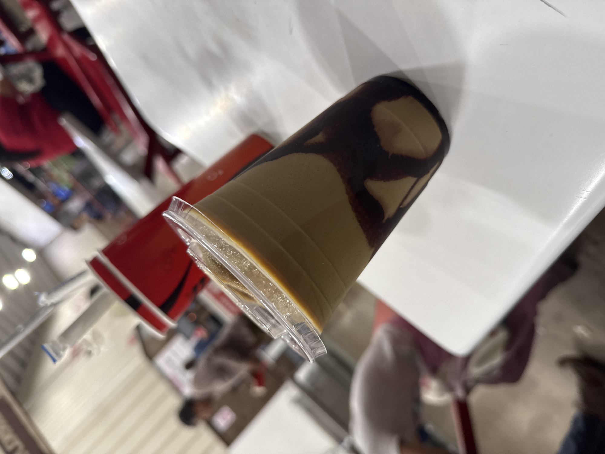 The image shows a drink with chocolate swirls on top, likely a mocha or similar beverage, sitting on a white table. Next to it is a red cup or container. The background suggests an indoor setting, possibly a cafeteria or food court. There are blurred figures of people and other objects in the background. The photo was taken at an angle, giving a slightly tilted perspective of the table and the objects on it. Information about Puebla and further detail is not possible from this information.