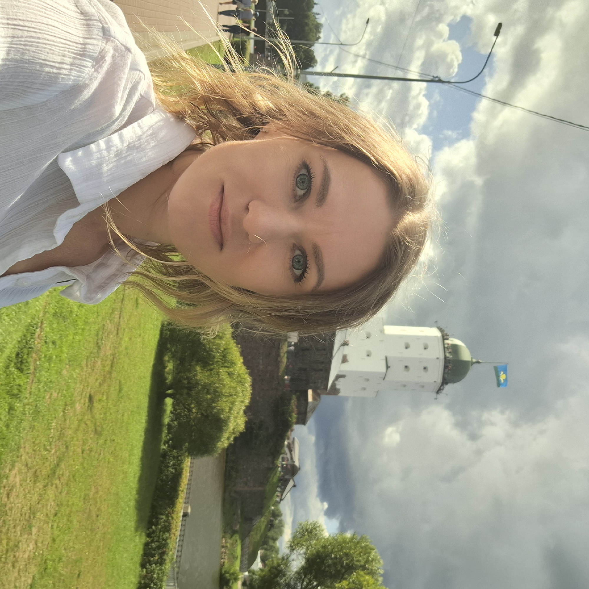 A woman with blonde hair and green eyes, wearing a white shirt, is taking a selfie outdoors. In the background, Vyborg Castle, a white fortress with a green-domed tower topped by a blue and green flag, is visible. The castle sits on a hill overlooking a body of water. The foreground shows a grassy area and a paved walkway with a lamppost. Several people are walking on the walkway in the distance. The sky is partly cloudy.