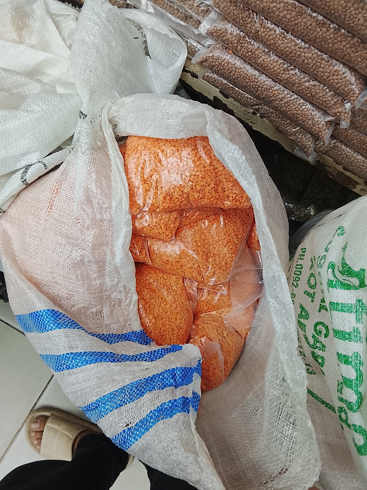 The image shows an open sack of red lentils in transparent plastic bags. Next to it are sacks of brown lentils. The sacks are in a store with tile floors. A person wearing a cream-colored slipper is visible. The location is Gulgasht Colony, Pakistan.
