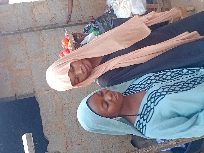 Two women in headscarves and traditional attire are positioned closely together. The woman at the top wears a peach-colored headscarf and a black outfit. She holds a tray of red fruits. The other woman, located below, has on a pale blue headscarf and a matching top with decorative trim. The background is slightly blurred, revealing part of a stone structure and other indistinct elements.