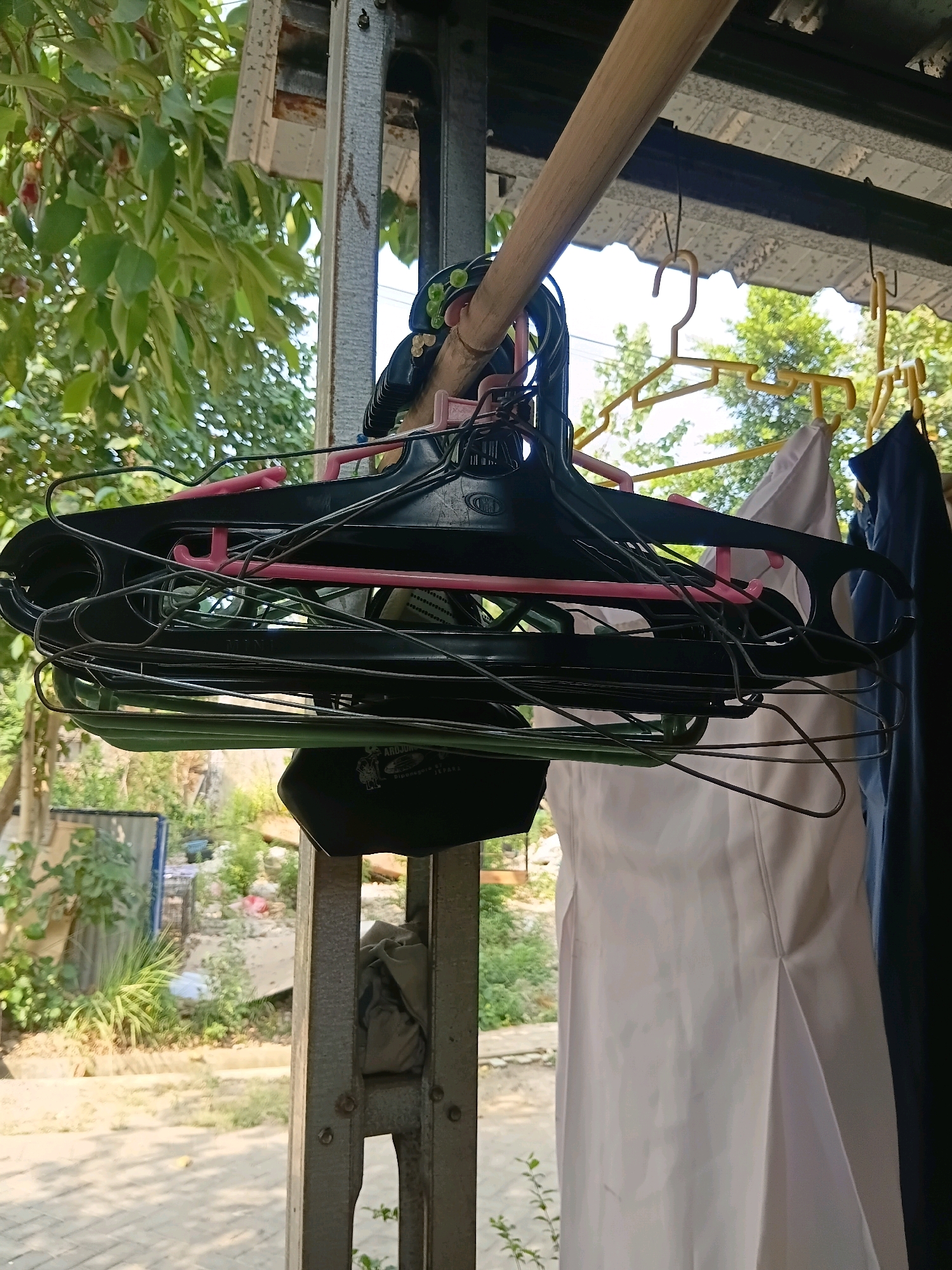 In the city of Cikal, Indonesia, a cluster of black and pink plastic hangers hangs from a wooden beam, laden with additional wire hangers. Among the hangers, a black cloth bag with white lettering is visible. To the right, white and dark blue garments hang on yellow plastic hangers, suspended from the same beam. The scene is set against an outdoor backdrop with lush green foliage and trees, suggesting a residential or communal area. The foreground shows a concrete pathway, with sunlight casting shadows. The overall impression is of an organized, outdoor drying area for clothing.