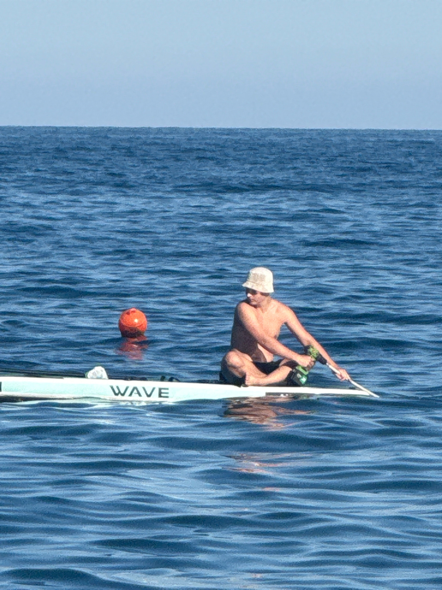 A man in a bucket hat and swim trunks sits cross-legged on a white paddleboard in the blue sea. He holds a paddle in his right hand and a green bottle in his left. To his left, an orange buoy floats in the water. The paddleboard has the word "WAVE" printed on its side. The sky is a pale blue, and the sea is calm with gentle ripples. The scene is set at the coast of Partenit, Ukraine.