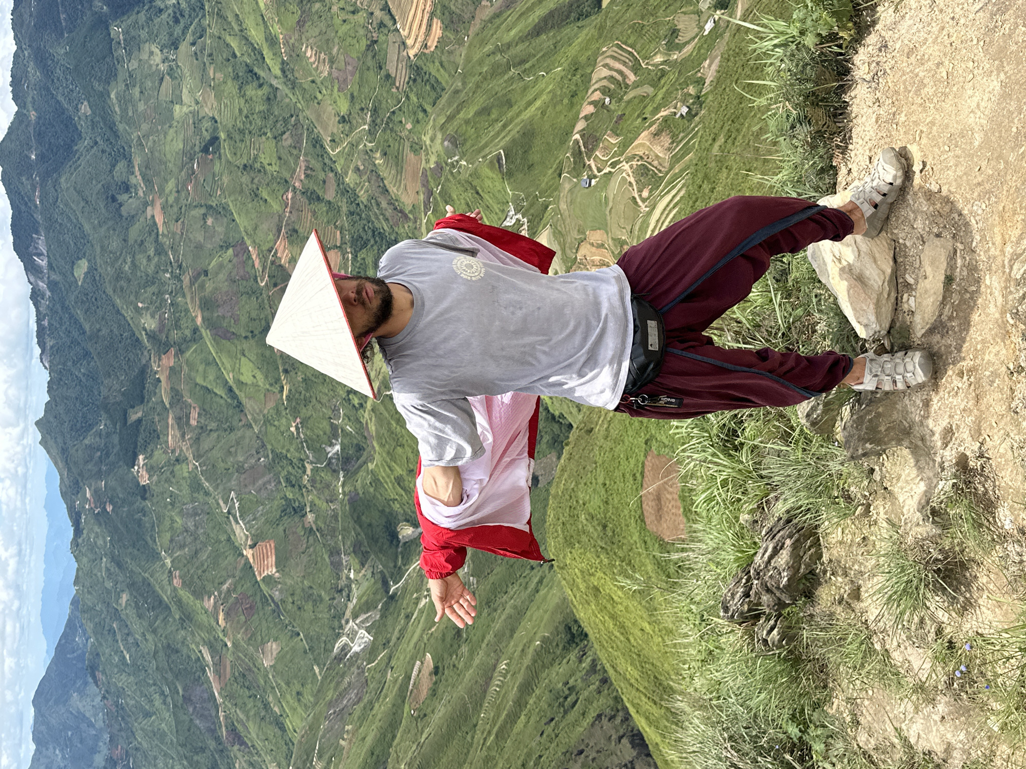 A person wearing a conical hat stands on a mountain slope in Ta Xua, Vietnam. The person is wearing a grey t-shirt, a red and white jacket draped over their shoulders, and dark red pants with blue trim. Their left arm is extended outwards, palm facing up. The background features steep, terraced hillsides covered in green vegetation and some brown patches, indicative of agricultural fields. The sky is partly cloudy. The ground in the foreground is rocky and covered in grass. The overall scene captures a landscape view from a high vantage point.