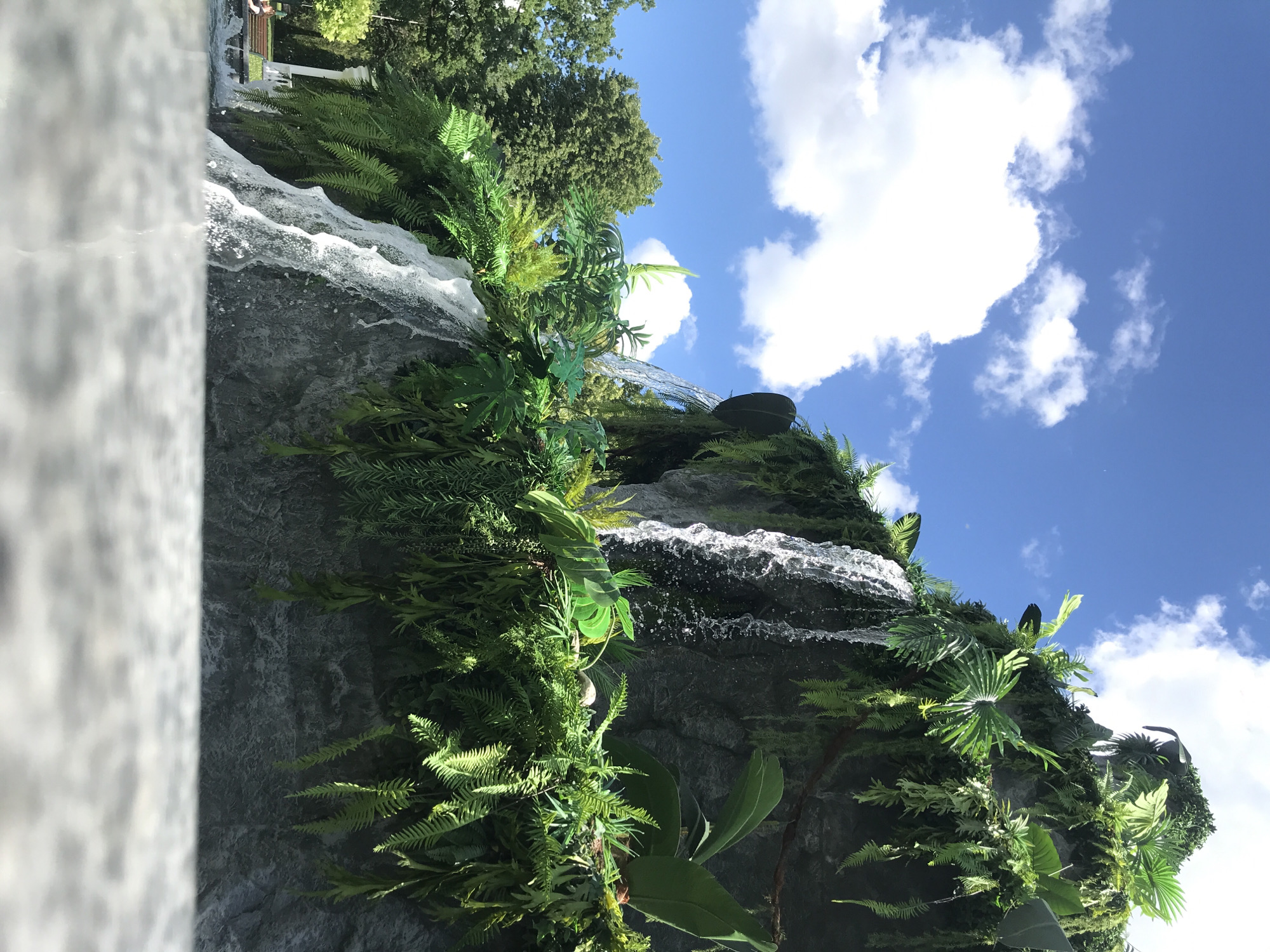 The image depicts a lush, artificial waterfall cascading over a rocky surface adorned with various green plants and ferns. The waterfall appears to be part of a man-made structure designed to mimic a natural rock formation. In the background, a clear blue sky with scattered white clouds provides a backdrop. To the left of the waterfall, a dense line of green trees is visible, suggesting a park or a natural setting. A white, classical-style structure, possibly a pavilion or a colonnade, is partially visible behind the trees. In the lower left corner, a portion of a park bench and a person are discernible, indicating a public outdoor space. The foreground is dominated by a blurred, textured surface, likely a stone ledge or a water feature's edge. The overall scene conveys a vibrant, tropical atmosphere within an urban park environment, specifically in Kharkiv, Ukraine, likely within the Kharkiv Zoo or a similar botanical garden attraction.