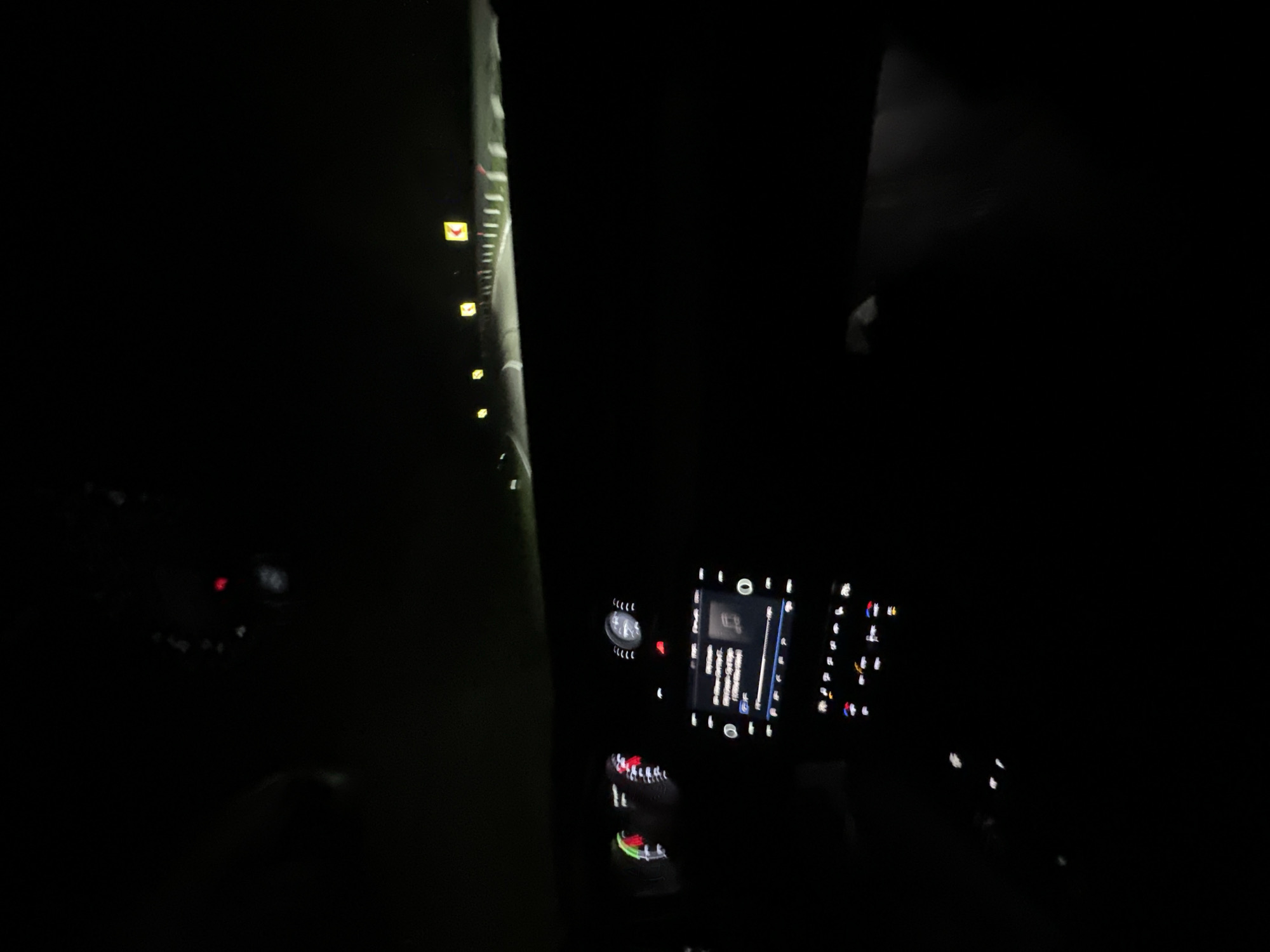 The image depicts the interior of a vehicle at night, viewed from the driver's perspective. The dashboard instruments, including a speedometer with a red needle, and an illuminated central infotainment screen displaying music information, are visible in the foreground. The vehicle's interior is largely dark, with light emanating from these displays. Through the windshield, a dark asphalt road is illuminated by the vehicle's headlights, receding into the distance. Multiple yellow square road signs with black chevron arrows, indicating a curve, are visible on the right side of the road, accompanied by white reflective road markers or a guardrail. The surrounding environment beyond the immediate illuminated road is obscured by darkness. No individuals are visible. The scene is located in Zaia, Bulgaria.