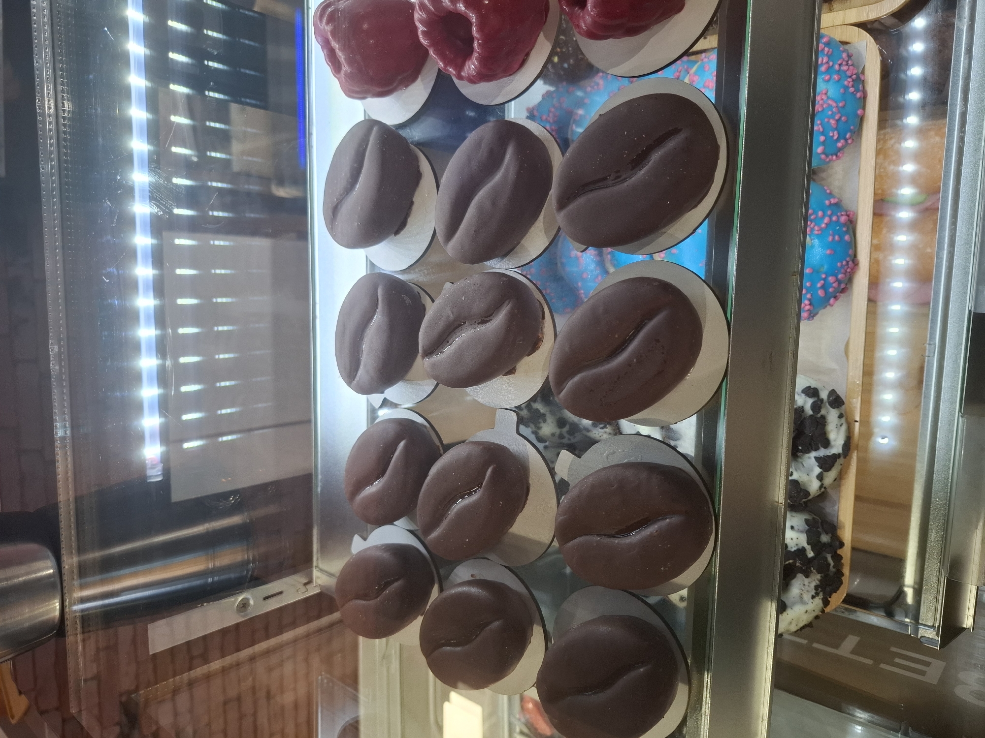 In Dubăsarii Vechi, Moldova, a refrigerated display case showcases an assortment of baked goods. The top shelf prominently features several chocolate-covered pastries shaped like coffee beans, each resting on a small white circular base. To the right of these are two vibrant red, ruffled pastries, possibly cakes or donuts. The lower shelf contains a selection of brightly colored donuts. Two have a blue glaze covered in small pink sprinkles, while others appear to have white icing with dark chocolate cookie crumbles. The display case is lit from above with bright LED lights, reflecting off the glass and the metallic interior. The background shows a brick wall and other bakery equipment.