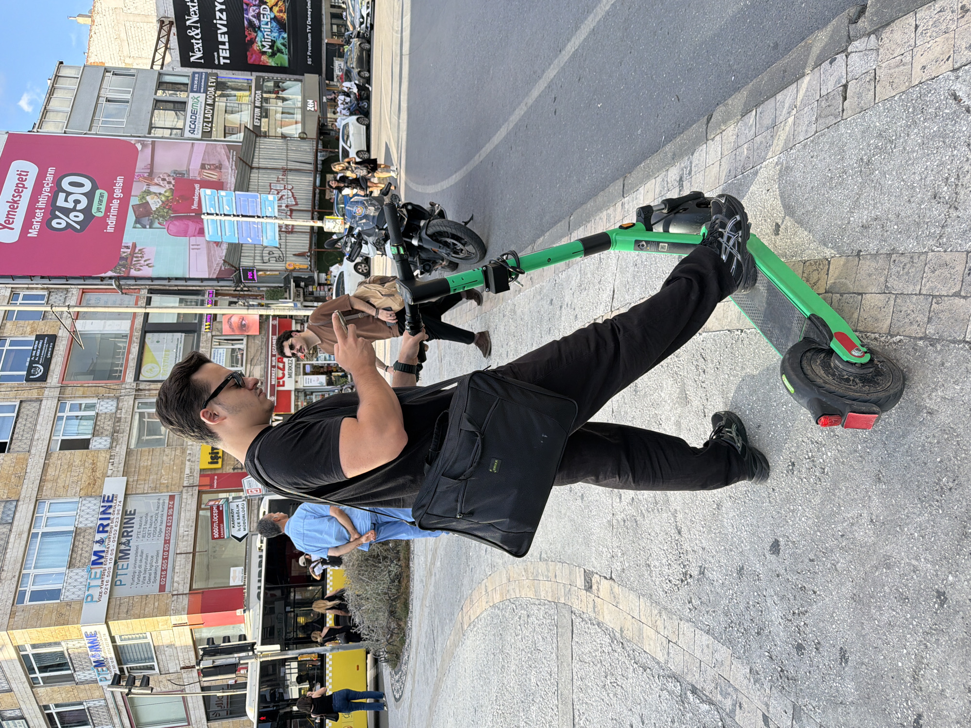 In Kadıköy, Turkey, a man stands on a sidewalk beside a bright green electric scooter, holding a smartphone. He is dressed in black, with black sunglasses and a black messenger bag slung over his shoulder. His left leg is on the scooter's deck, and his right leg is bent, appearing to be in the process of mounting or dismounting. Behind him, several people are visible, including a man in a blue shirt and a woman in a brown top. A yellow bus and a motorcycle are present in the background, along with numerous building facades displaying various advertisements. The ground is paved with light-colored concrete and decorative stone, and a curb separates the sidewalk from a paved road. The scene is set on a street with moderate pedestrian and vehicle traffic under a clear sky.