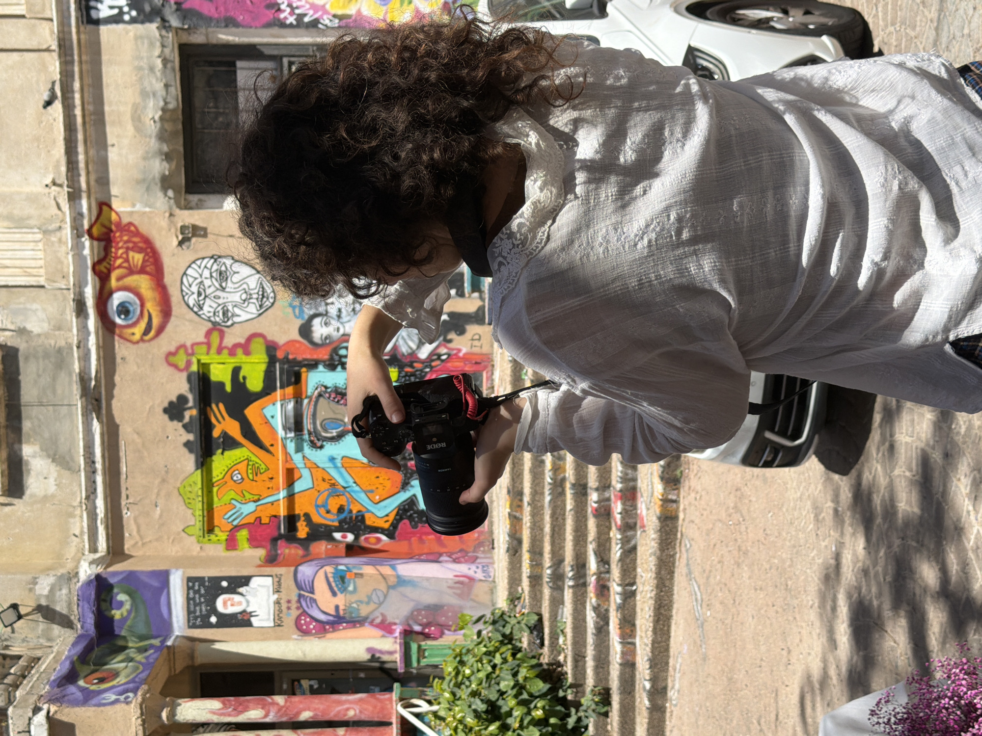 A person with curly dark hair, seen from behind and the side, is photographed holding a black camera with a large lens in an outdoor urban setting in Tel-Aviv, Israel. They are wearing a textured white shirt with lace detailing on the collar. The camera has "RØDE" and "TAMRON" visible on its components.
The background features a vibrant wall extensively covered in graffiti. Notable artwork includes a large red and gold fish with a prominent blue eye, a white abstract face drawing, colorful abstract human-like figures, a green and purple creature, and a stylized face with blue eyes and purple hair. Visible text within the graffiti includes "Kamoach Kuchi" and "P!".
The scene is brightly lit by natural sunlight, casting distinct shadows, suggesting a clear, sunny day. Part of a white car is visible in the upper right. On the lower right, there is a small white table adorned with purple flowers, and green leafy plants grow near the base of the wall. Ribbed concrete steps or a raised pathway run diagonally across the middle-right of the frame. The person is actively engaged in taking a photograph.