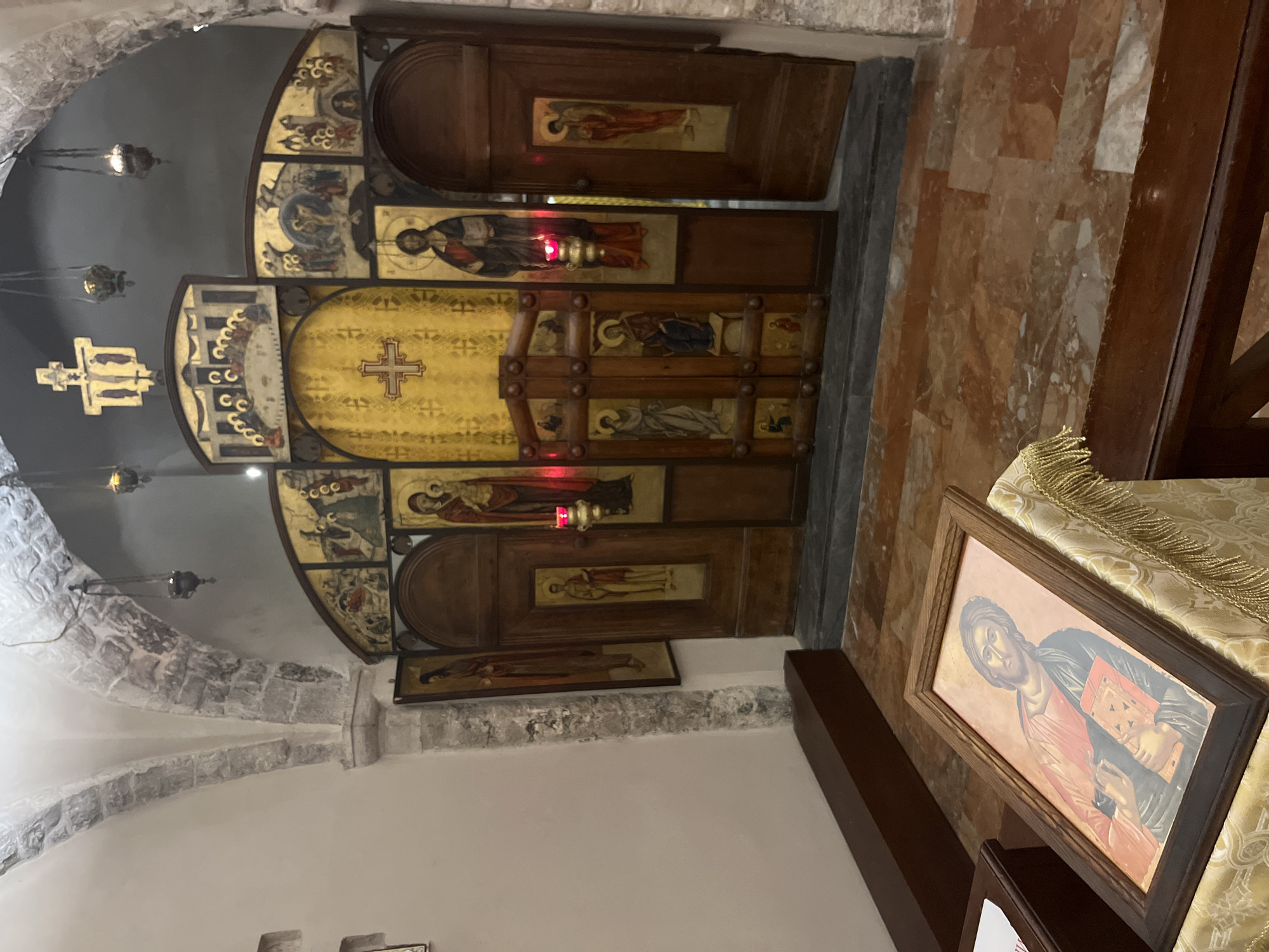 This is a photograph of the iconostasis in the Basilica di San Nicola in Bari, Italy. The iconostasis is a wall of icons and religious imagery that separates the sanctuary from the nave of the church. The central part of the iconostasis features large wooden doors, intricately carved and adorned with religious paintings, known as icons. These icons depict various figures from Christian tradition, including Jesus Christ, the Virgin Mary, and saints.
The iconostasis is illuminated by a combination of natural light filtering in from unseen sources and artificial lighting, including hanging lamps and the warm glow of candles placed before some of the icons. The architectural setting includes stone arches and a vaulted ceiling, suggesting an ancient and sacred space.
In the foreground, on a draped table, rests a framed icon of Jesus Christ. The floor is paved with marble tiles in shades of red and white. The overall impression is one of reverence and historical significance, characteristic of a major religious site in Italy.