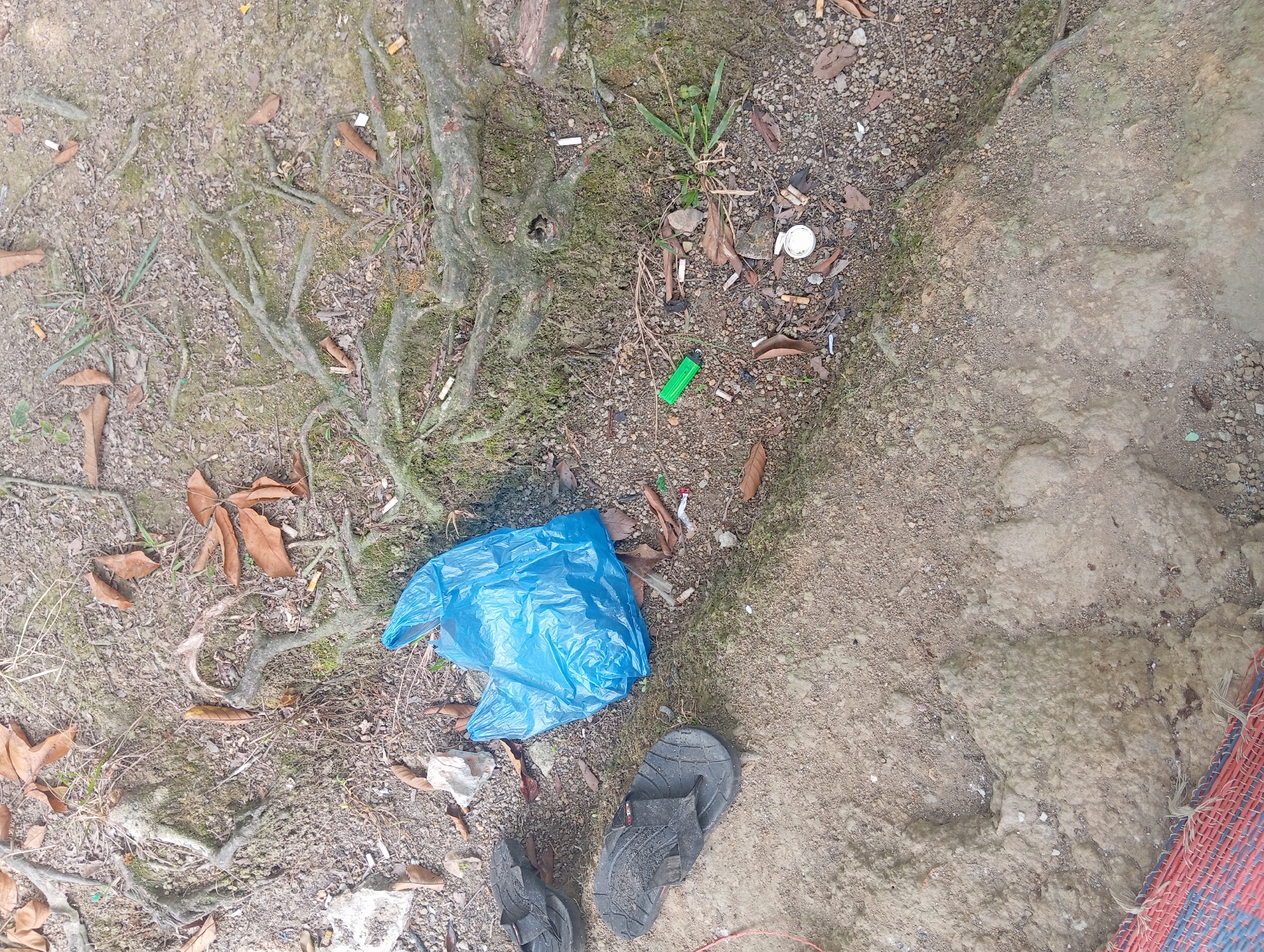 An outdoor scene in Rejosari, Indonesia, shows ground litter and personal items. A bright blue plastic bag lies open on the dirt and root-covered ground. To the left of the bag are two black flip-flops. Scattered on the ground are numerous cigarette butts, dried leaves, small stones, and a green lighter. The area appears to be a pathway or natural space with exposed tree roots and uneven terrain. A woven mat with red and blue stripes is visible in the lower left corner.