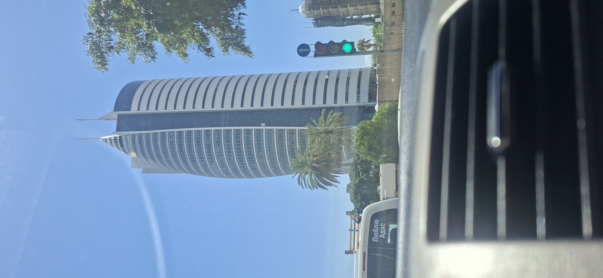 A tall, modern, curved office building dominates the center of the frame. It features a glass facade with horizontal white bands. The building has two pointed spires at its apex. In front of the building, there are two palm trees. To the right of the building, a traffic light is visible with the green light illuminated. Further to the right, another building under construction is present. In the foreground, the interior of a vehicle is visible, with a dashboard and air vent in focus. A white van with text in Cyrillic script reading "Люблю Адас" is parked to the left of the main building. The sky is clear and blue. The image appears to be taken during the daytime.