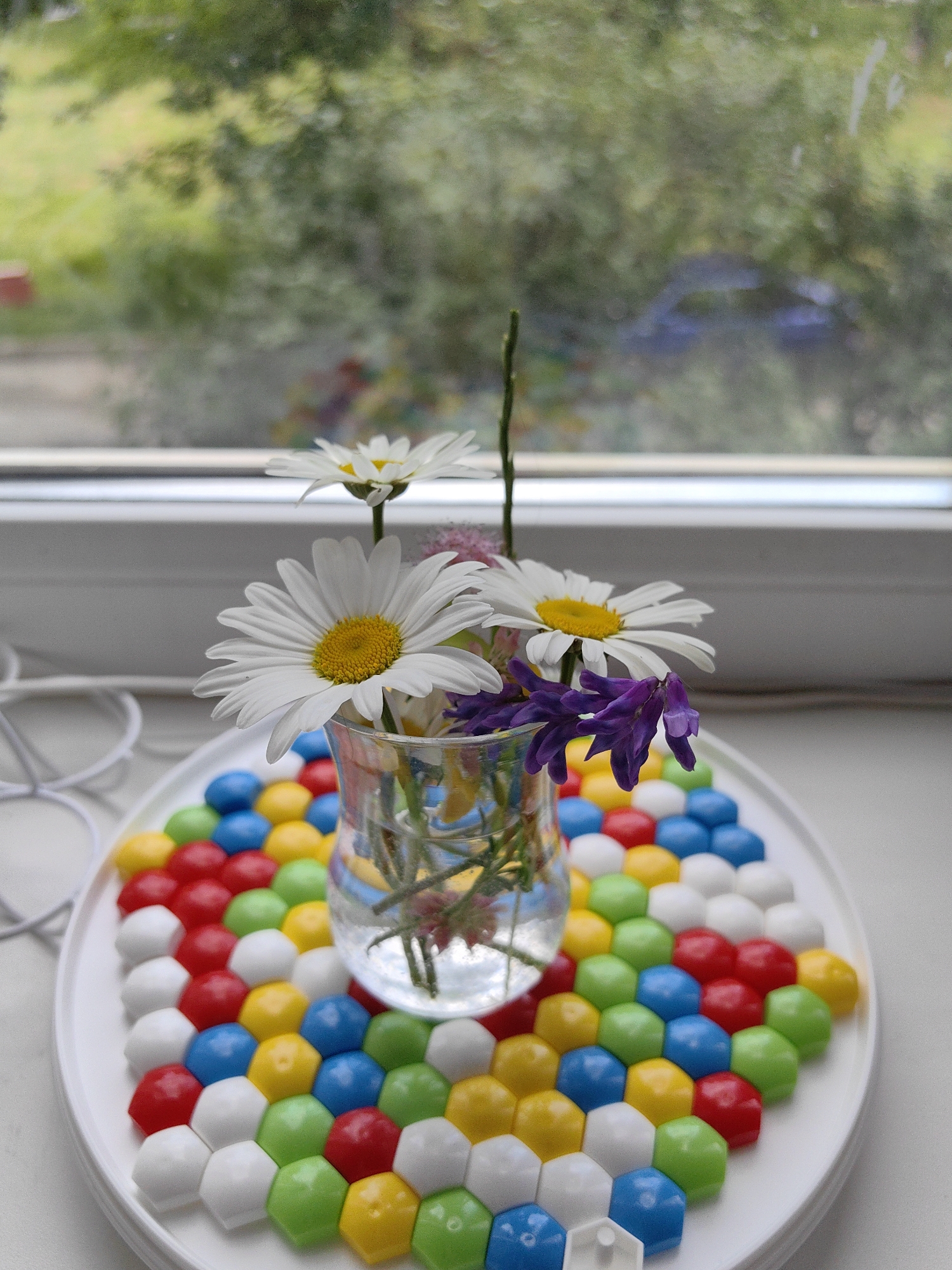 A still life arrangement features a clear glass vase containing white daisies and purple flowers, placed on a white circular tray filled with colorful hexagonal mosaic tiles. The tray is positioned on a windowsill with a blurry green and yellow background suggesting foliage outside. The lighting is natural, coming from the window. The overall scene is indoors, with the windowsill serving as the primary surface for the objects. There are no people or animals present. The background details are indistinct, but the focus is on the flowers and the mosaic tray.
