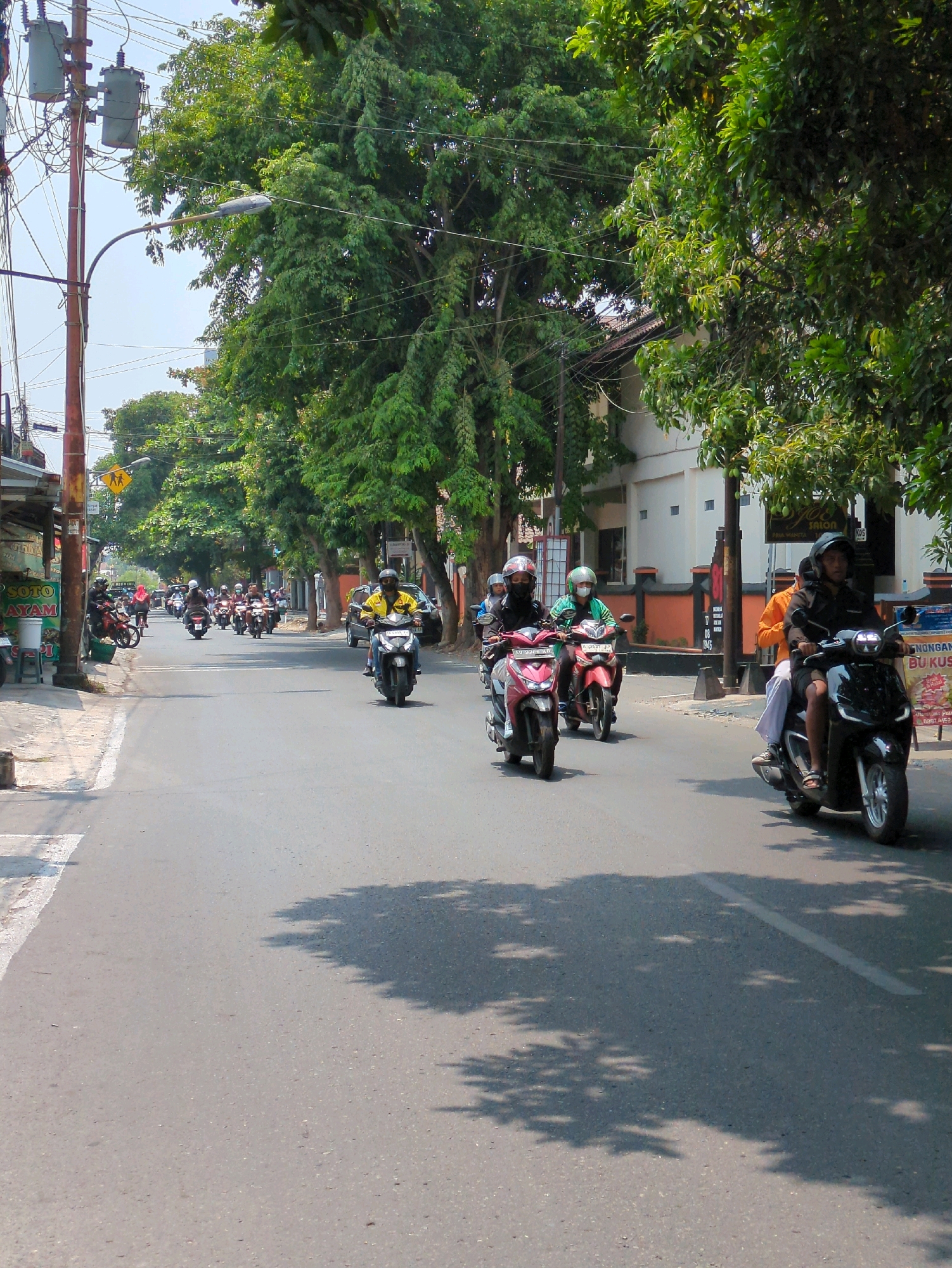 The image depicts a street scene in the city of Surakarta, Indonesia, characterized by a procession of motorbikes on a paved road lined with trees and buildings. The perspective is from street level, looking down the road as traffic moves away from the viewer.
In the foreground, a dark-colored motorbike is being ridden by a person wearing a helmet and an orange shirt. They are in the right lane of the road. To their left, and slightly behind, are two more motorbikes. The first, a pink motorbike, is ridden by a person in a green helmet and a green shirt. The second, a red motorbike, is ridden by a person wearing a black helmet and a black jacket. These motorbikes are also in motion.
Further down the road, a larger convoy of motorbikes is visible. Most of the riders are wearing helmets. The traffic appears to be moving in the same direction. One motorbike in the middle distance has a rider in a yellow shirt and helmet.
The environment suggests a daytime scene under clear skies. The road surface is asphalt, with a white line marking the lane. Shadows of trees are cast across the road, indicating the presence of dense foliage. On the left side of the road, there are buildings, some appearing to be shops or small businesses, with awnings and signage. One sign visible on the left has text that includes "SOTO AYAM". On the right side of the road, there are more substantial buildings, one with a white facade and an orange trim, and trees providing shade. Utility poles with electrical wires are visible along the side of the road, typical of urban infrastructure. A street lamp is mounted on a pole over the road.
The overall impression is one of typical urban traffic flow in Indonesia, with a strong reliance on motorcycles for personal transportation.
