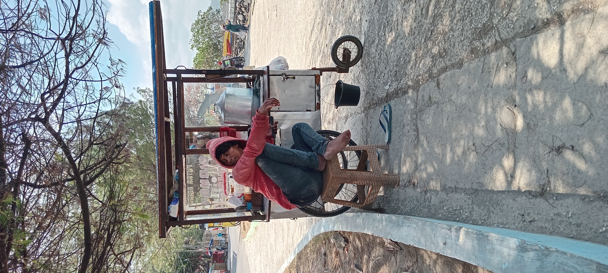 A man wearing a red hooded sweatshirt and blue jeans sits on a brown plastic stool next to a food cart in Desa Pagandaran, Indonesia. The man is leaning against the cart, with his legs crossed and his feet bare. His left hand is resting on his lap, and his right arm is extended, with his hand on the cart. A pair of blue and white flip-flops lies on the ground next to the stool. The food cart is made of metal with a wooden frame and a blue tarp roof. It has a large stainless steel pot inside. A black bucket hangs from the side of the cart. The ground is paved with concrete and dirt. In the background, there is a road with vehicles and people, as well as trees and a body of water. The sky is partly cloudy.