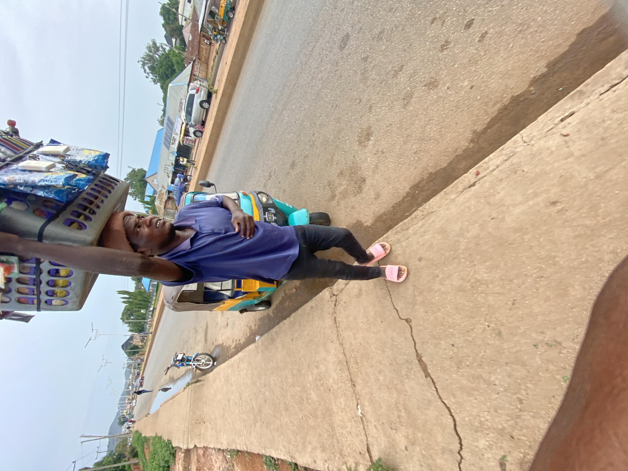 The image shows a man in Bukuru, Nigeria, carrying a large load on his head. He wears a purple shirt, black pants, and pink sandals. He is walking on a sidewalk next to a street with vehicles, including a light blue and yellow three-wheeled vehicle. The background includes buildings, trees, and power lines. The road is paved, and the sidewalk is concrete.