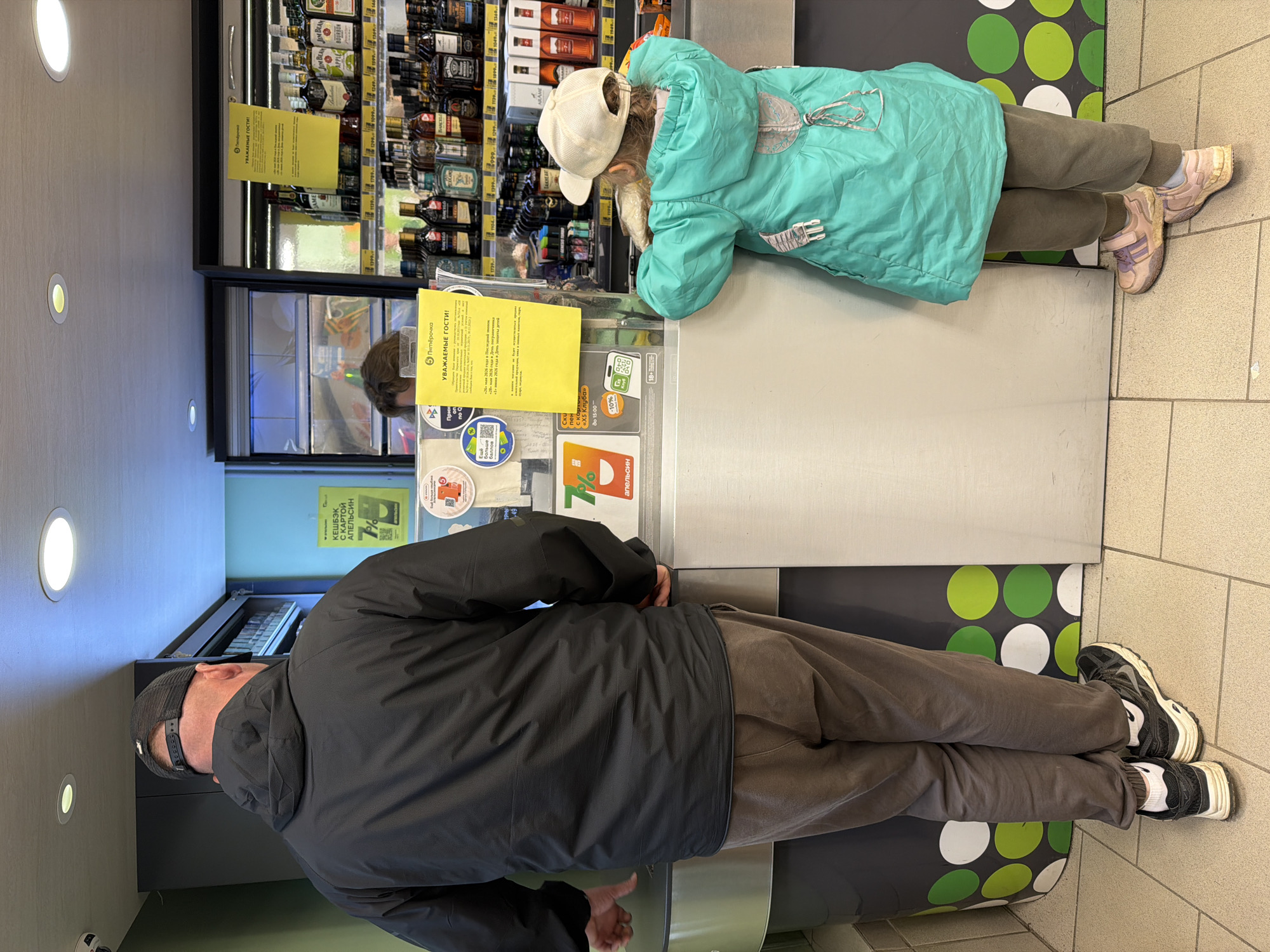 A father and daughter are standing at a checkout counter in a grocery store in Chaikovsky, Russia. The father, wearing a black jacket, brown pants, and a black baseball cap, stands in line. The daughter, wearing a light blue jacket, grey pants, a white baseball cap, and pink sneakers, leans on the counter, looking at the items displayed behind the counter. These items are mostly bottles of liquor. The counter is grey, and the floor has green and white polka dots near the father. The walls are light in color. Yellow signs with Russian text are visible on the counter and near the liquor bottles.