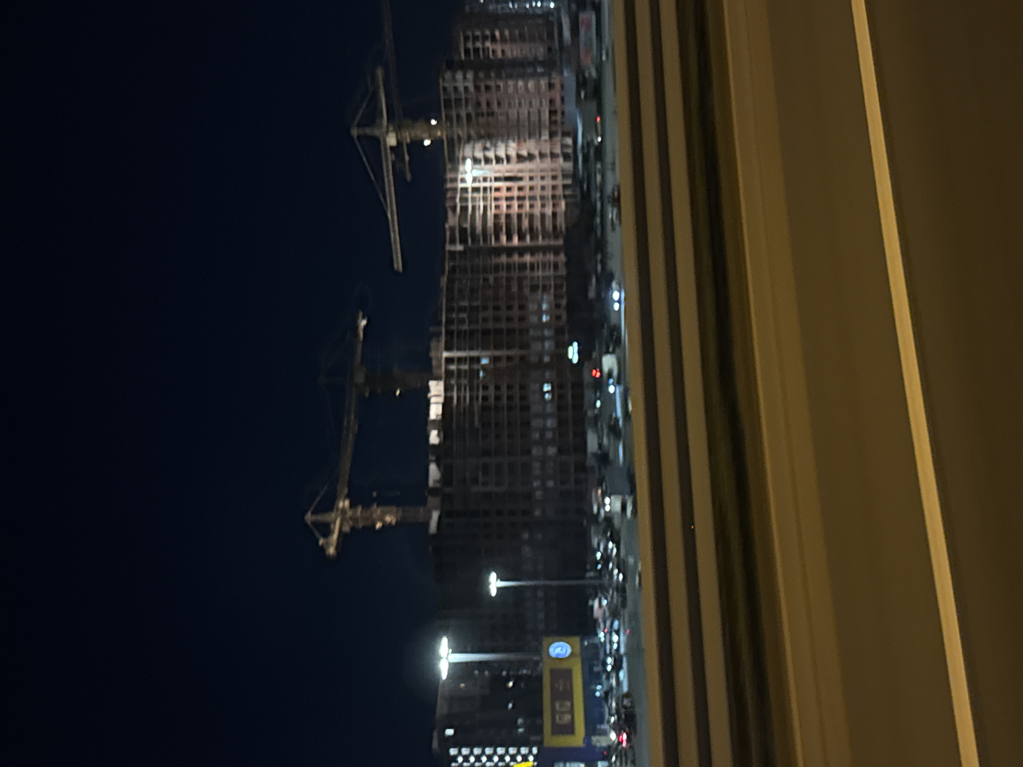 This nighttime image depicts a construction site in Saint Petersburg, Russia, featuring large cranes and an unfinished building. The building appears to be a multi-story residential complex, with its concrete framework exposed. Several vehicles are visible in the foreground, suggesting a nearby road or parking lot. The lighting is dim, with streetlights and lights from the construction site illuminating the scene. The overall atmosphere is one of ongoing development and urban growth.