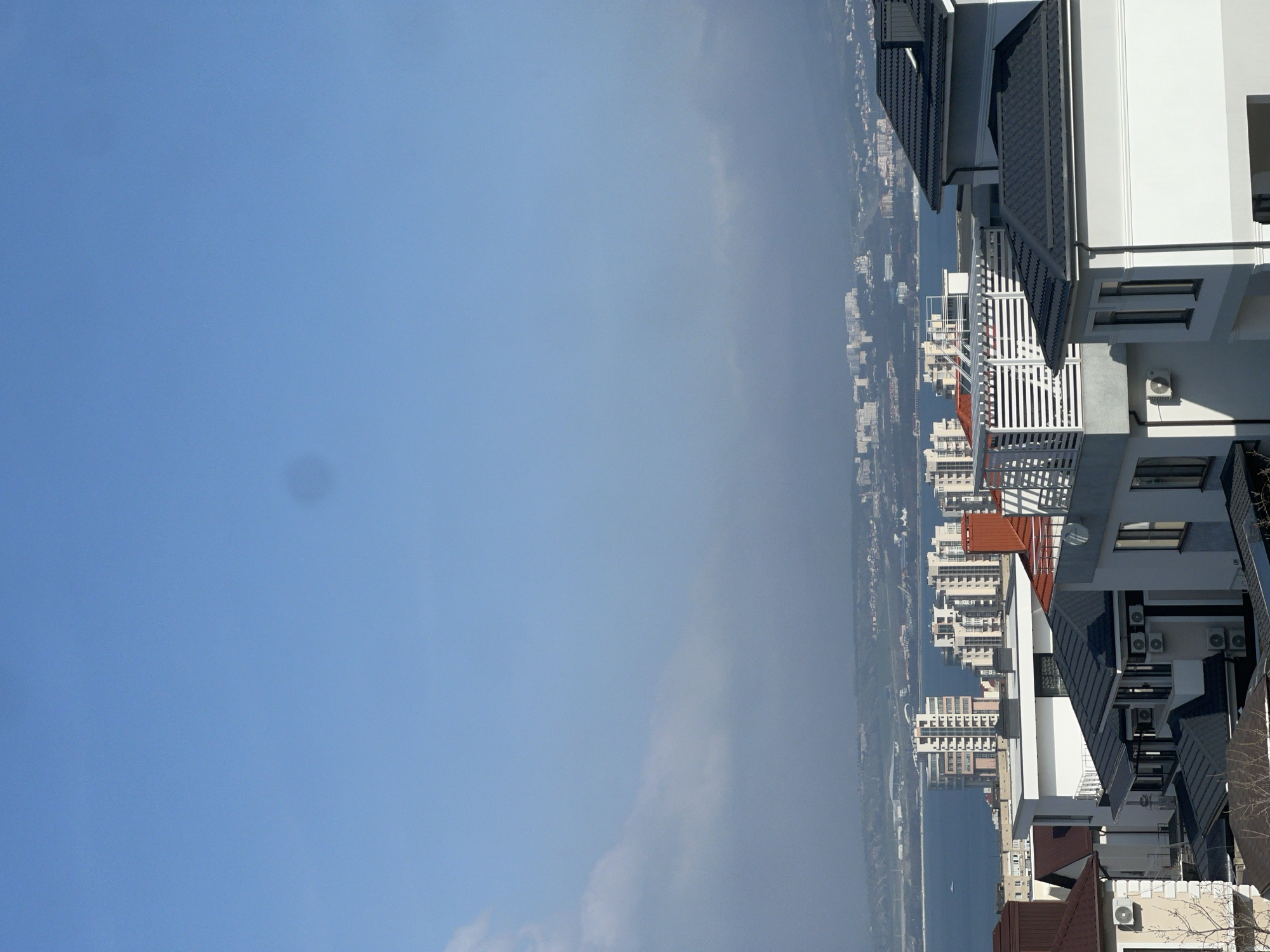 The image shows a view from a high-rise building in Gelendzhik, Russia. The sky is predominantly light blue. Below, there is a coastal cityscape, with buildings stretching along the coastline. In the foreground are rooftops and upper floors of modern buildings with white facades, dark roofs, and multiple air conditioning units. The buildings show a mix of architectural styles with features like balconies with white railings and angled rooflines. The overall impression is a modern urban landscape with a coastal setting.