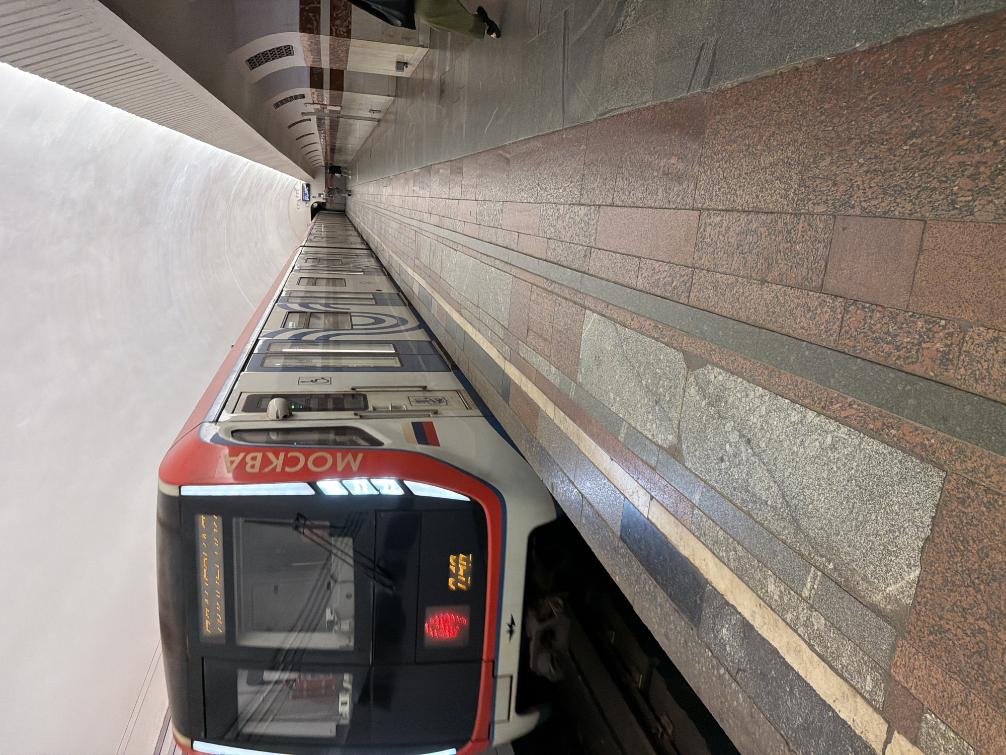This image shows a train in a subway station in Moscow, Russia. The train is red and white with the word "МОСКВА" written vertically on the side. The destination board on the front of the train displays text in Cyrillic letters, which appears to be "АЛЕКСАНДРОВСКИЙ САД 1140". The station has a white, arched ceiling and a platform tiled with granite in shades of grey, pink, and red. A person can be seen walking on the platform in the background. The lighting is soft and diffused, typical of an underground station.