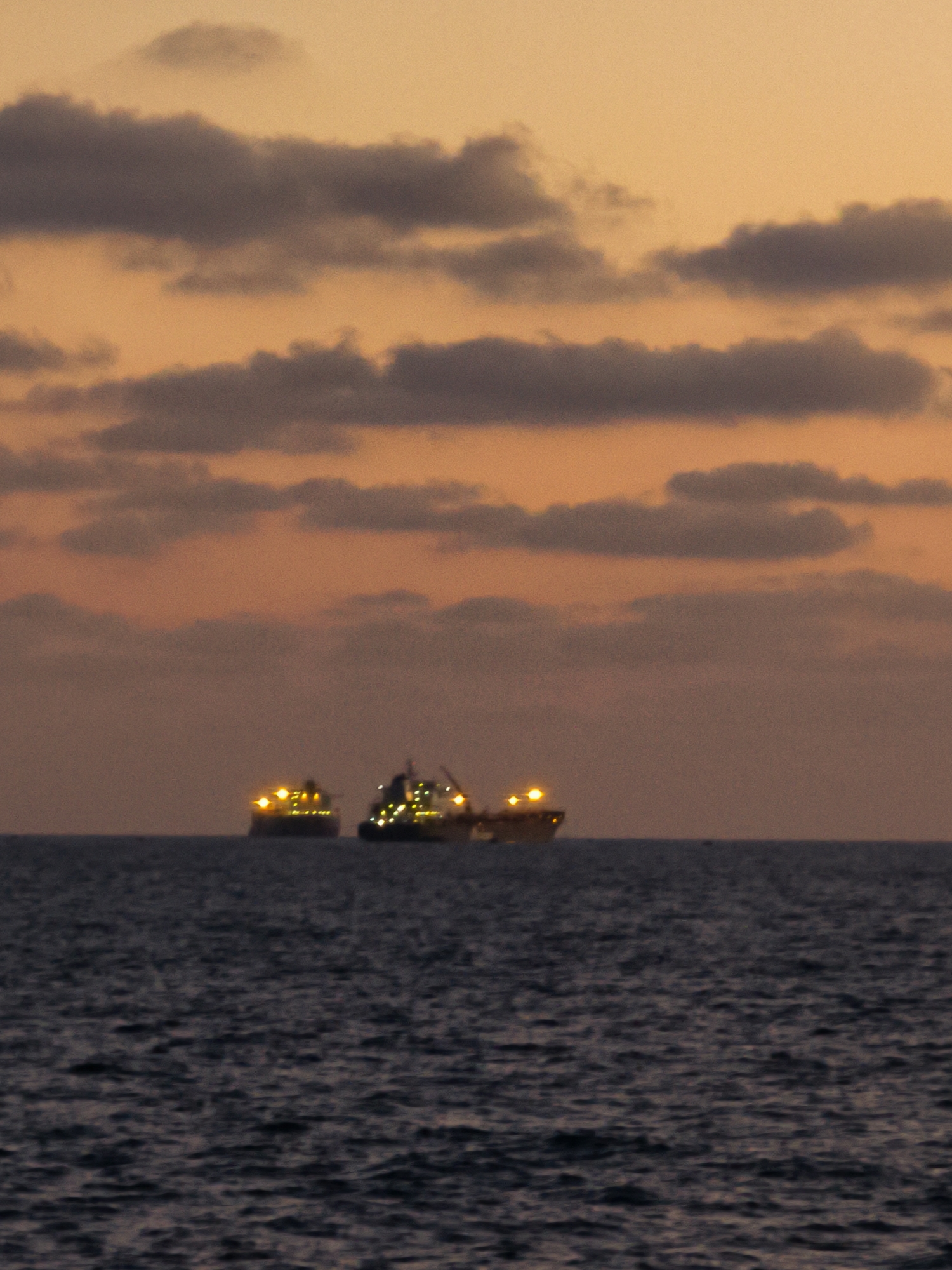 Two cargo ships are visible on the water in the distance, their lights illuminated against the dusky sky. The sky is filled with clouds, tinged with the orange and pink hues of sunset. The sea is dark and appears somewhat choppy. The image likely depicts a scene off the coast of Ashkelon, Israel, given the description. There are no people visible in the image.