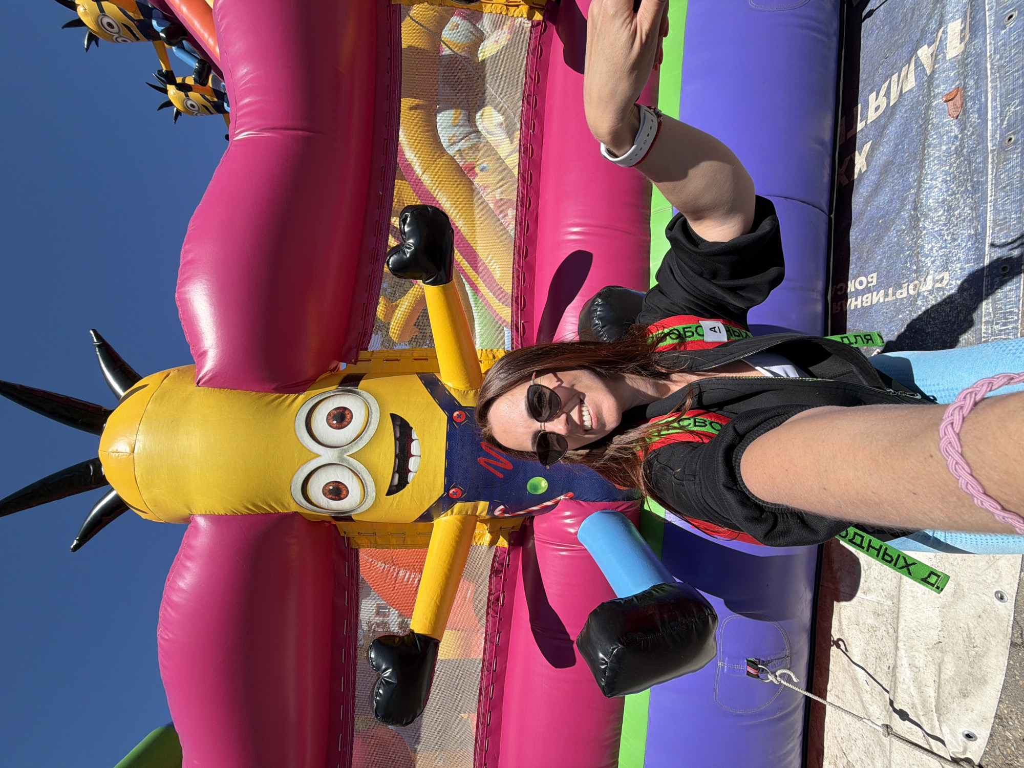 The image captures a woman taking a selfie at an outdoor recreational area in Birobidzhan, Russia, on a bright, sunny day. She is smiling broadly and wearing sunglasses, a black jacket, and a white wristband, with a pink braided bracelet on her right wrist.
The scene is dominated by a vibrant, inflatable structure, likely a bouncy castle or obstacle course, featuring prominent sections in pink, purple, yellow, blue, and green. To the woman's left stands a large, cheerful inflatable Minion character, with its distinctive yellow body, large goggles, and spiky black hair. Smaller Minion-like figures are also visible in the upper background against a clear blue sky, suggesting a larger themed display. A blue support element is visible near the woman's body, indicating she is interacting closely with the inflatable.
The ground beneath the inflatable is covered with a white or light-colored tarp, which is secured by ropes and anchors. The strong sunlight casts clear shadows, indicating it is likely midday or early afternoon.
Reversed Cyrillic text is visible on various elements. On the purple section of the inflatable, phrases like "СПОРТІВНЫЙ БОКСА" (Sporting Boxing) can be partially read. Another green strap or tape near the woman's right arm also bears reversed Cyrillic text. Further reversed text, including "ЛАМЯТ" and parts of "СПОРТІВНЫЙ БОКСА," are visible on the white tarp covering the ground, suggesting it might be a repurposed banner. The overall impression is one of fun and leisure at an outdoor event.