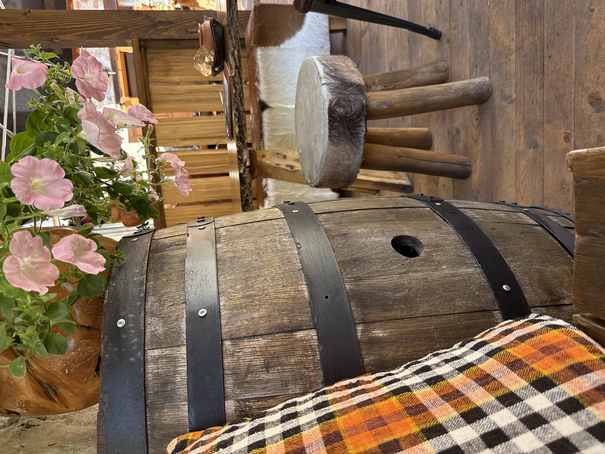 The image depicts an outdoor seating area with rustic wooden furniture and decorative elements. In the foreground, a portion of a weathered wooden barrel is visible, adorned with black metal bands. A plaid pillow, featuring orange, black, and white checks, rests against the barrel.
Behind the barrel, a small, round stool with a fur-covered seat and three wooden legs stands on a wooden floor. Further back, a wooden table with a natural wood edge is set with a small, dark brown bowl containing a golden object. A rustic wooden fence forms a backdrop behind the table.
Hanging above the table are pink petunias in bloom, cascading from a wooden planter. In the mid-ground, benches with white and brown cowhide upholstery are visible. The overall atmosphere suggests a cozy, rural, or garden setting, possibly in Shiroka Laka, Bulgaria, known for its traditional architecture and natural beauty. No people are present in the image.