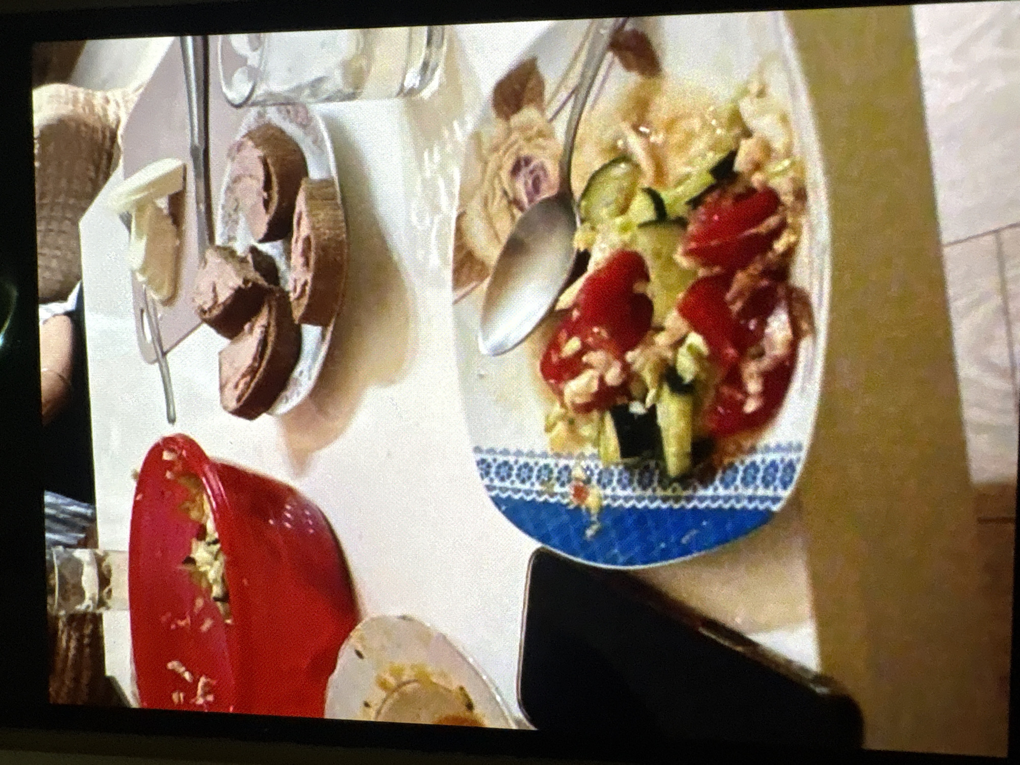 A casual meal is taking place at a table, likely in a home setting, in Batumi, Georgia. The image captures a close-up view of food items arranged on a white tablecloth.
In the foreground, a plate with a salad is visible, featuring chopped tomatoes, cucumbers, and what appears to be some sort of grain or pasta. A spoon rests on the plate, suggesting the meal is in progress. To the left, a black smartphone is placed on the table.
Further back, a red bowl contains remnants of a dish, possibly a salad or appetizer. Adjacent to it, a plate holds several pieces of bread topped with a spread, resembling pâté or cheese. A glass of water is also present on the table. In the background, out of focus, a woven basket and another glass are visible. The overall lighting suggests an indoor setting, possibly during daylight hours, given the ambient light illuminating the scene.