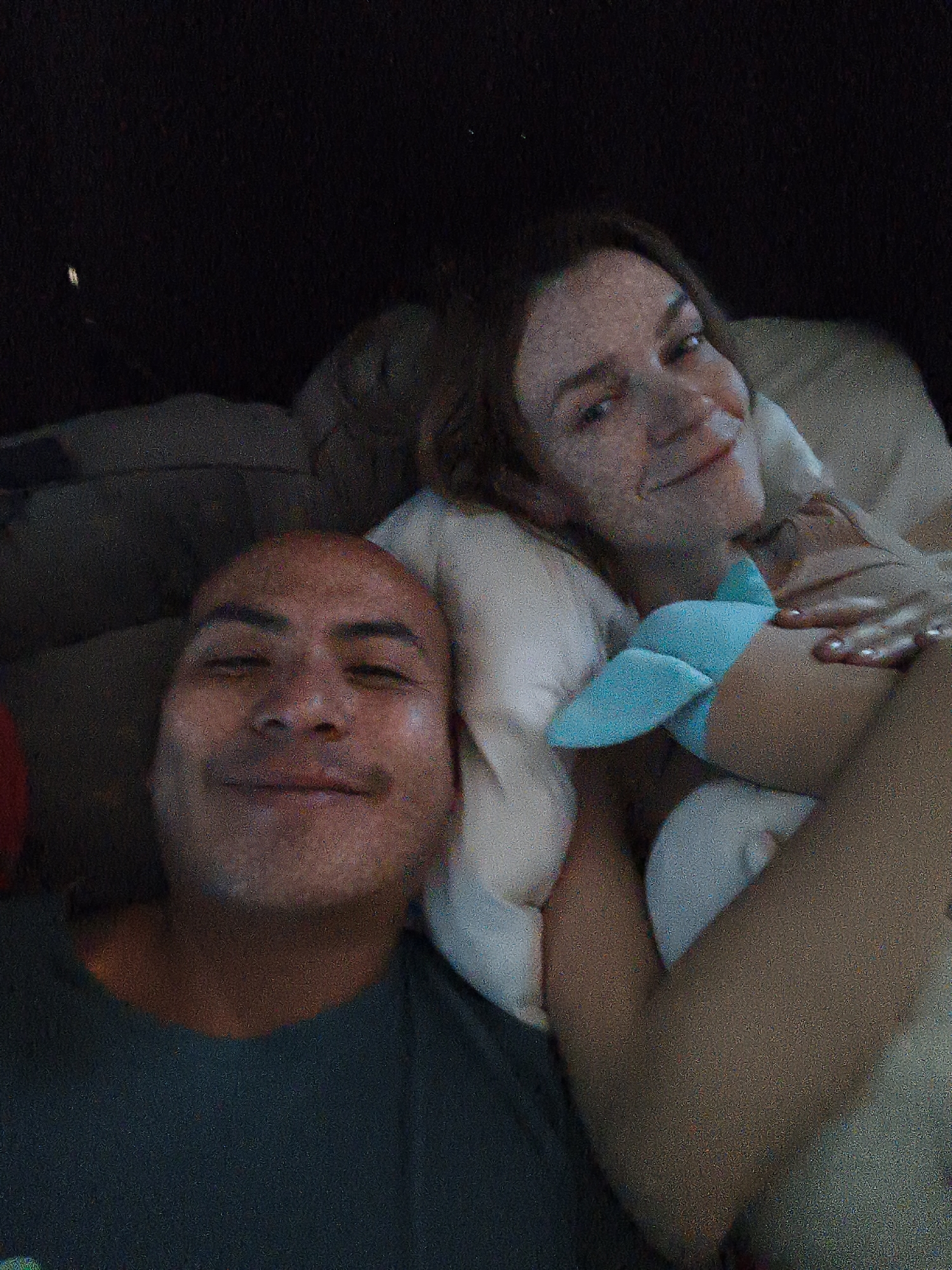 A low-angle shot depicts two individuals lying in bed. On the left is a bald man, positioned closer to the camera, wearing a dark-colored t-shirt. To the right is a woman with light skin and light brown hair. She is nestled against a white pillow and holding a light blue stuffed animal. The scene is indoors, presumably in a bedroom, with the background appearing dark.