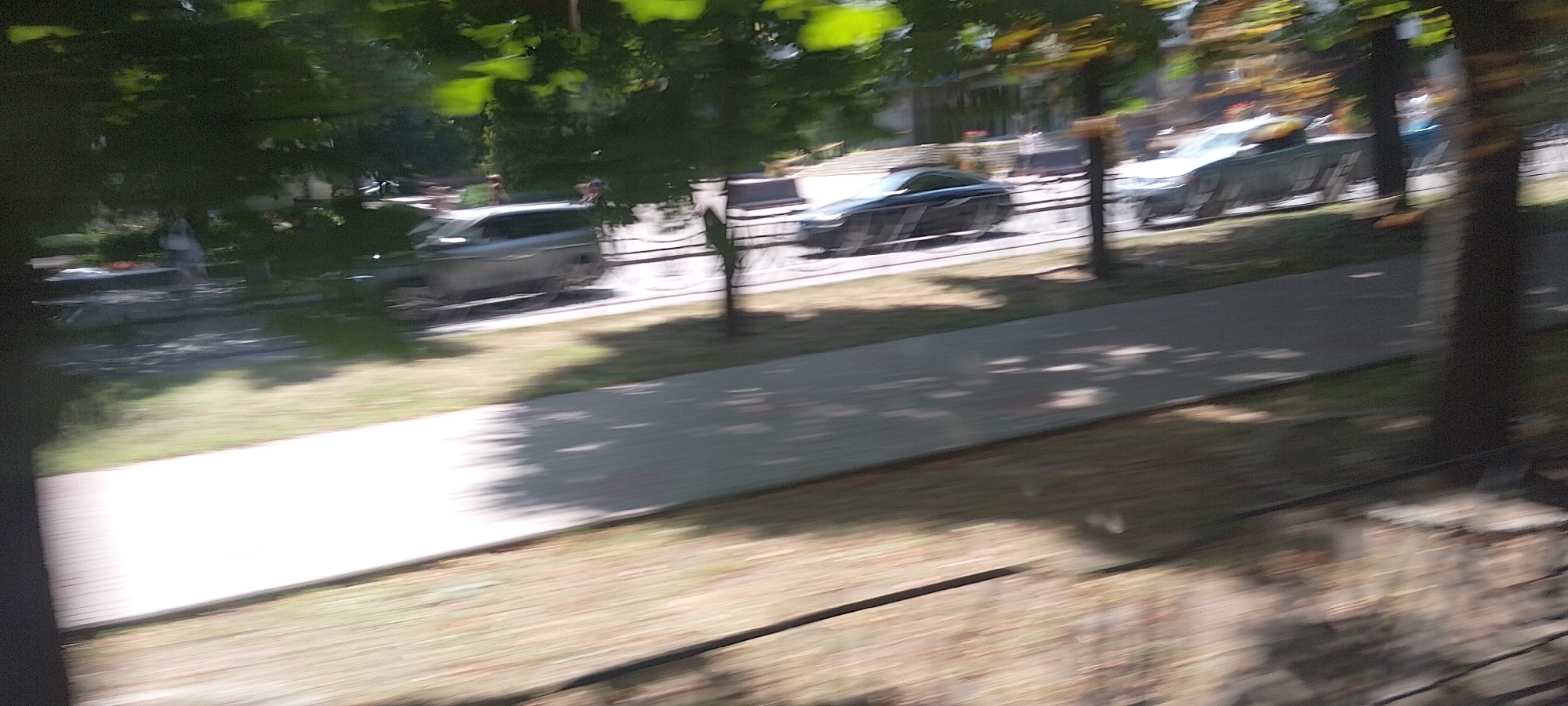 The image depicts a blurred outdoor scene, likely taken from a moving vehicle, showing a paved pathway, grass, trees, and several vehicles on a road in the distance. A silver SUV is visible on the road, as is a dark sedan. There are also indications of people standing near a table in the background. The overall impression is one of motion and a typical urban park or street environment. The reference to the city of Cherkasy, Ukraine, suggests this scene is located there, potentially along one of the city's roads or park areas with pedestrian walkways. The time of day appears to be daytime, with sunlight casting shadows through the trees.