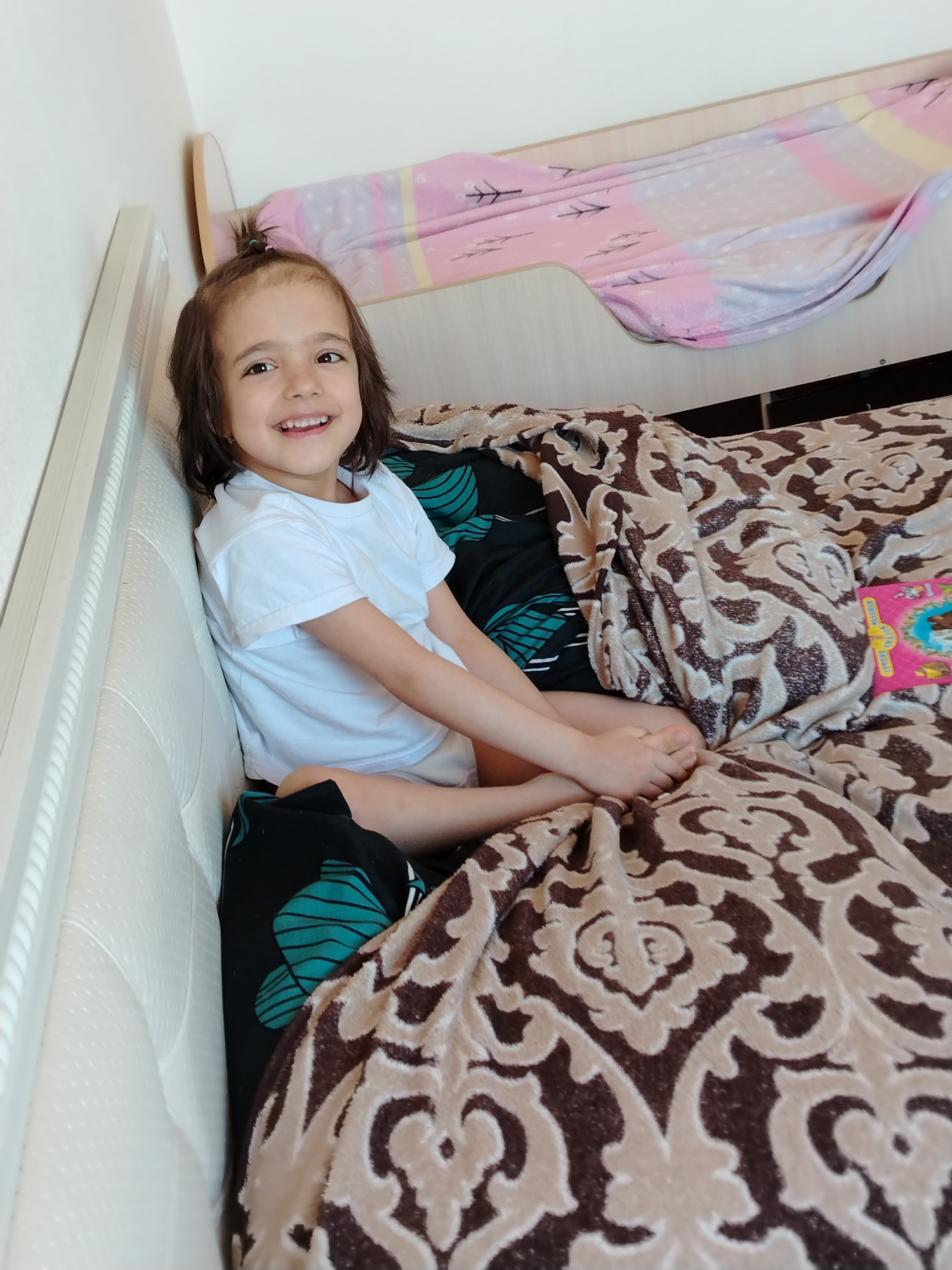 A smiling child, likely female, is seated on a bed. The child has brown hair with a small ponytail on top, brown eyes, and is wearing a white t-shirt. The bed has a white headboard, and is covered with a brown and beige patterned blanket. There is a black and teal patterned sheet partially visible under the blanket. A pink object, possibly a book or toy, is partially visible on the bed as well. There is a pink and white patterned blanket over a bunk bed safety rail in the background. The location is possibly an apartment interior in Noviy, Russia, based on the user's query, but there are no identifying external environmental details in the image to confirm. The child is interacting with the camera, appearing happy and engaged.