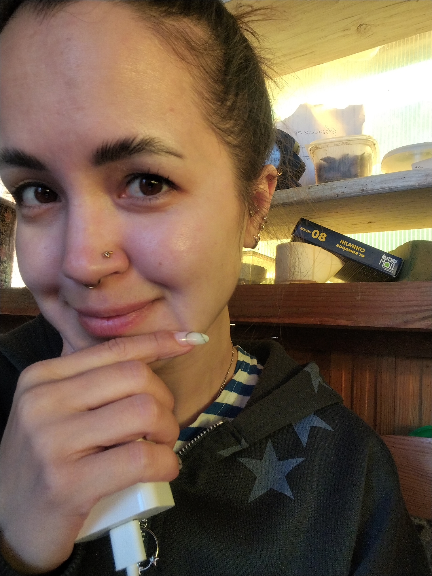 A woman with dark eyebrows and brown eyes is holding a white object in her hand and resting her chin on her finger. She has multiple earrings in her left ear and two nose piercings. She is wearing a striped shirt underneath a dark gray hooded sweatshirt with light blue stars. Behind her, a wooden shelf is visible with various containers and a box with Russian writing. The writing on the box says "СУСТАВИНОВ-КОМПЛЕКС". The city of Noviy, Russia is not identifiable in the image.
