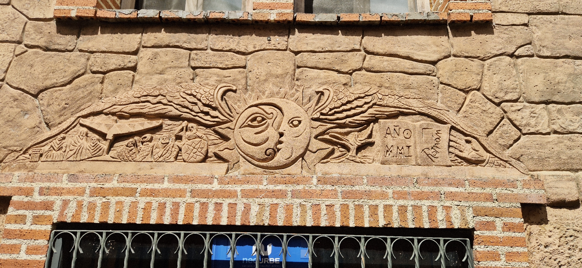 The image shows a stone carving above a window. The carving features a sun and moon face, with wings extending outwards. To the left of the sun and moon, there are figures that appear to be people or deities, some wearing robes and others with elaborate headdresses. A triangular structure is also depicted above these figures. To the right of the sun and moon, there are carved inscriptions that appear to be in Roman numerals and possibly a name. The carving is set against a background of rough, irregularly shaped stones, with a brick facade below. The lighting suggests it is daytime, with shadows indicating sunlight hitting the relief. The overall style of the carving is artistic and possibly symbolic, with the sun and moon motif suggesting themes of duality or celestial cycles. The inscription "MMI" likely refers to the year 2001, providing a date for the artwork. The word "Román" is also visible. The scene is architectural, part of a building's facade.